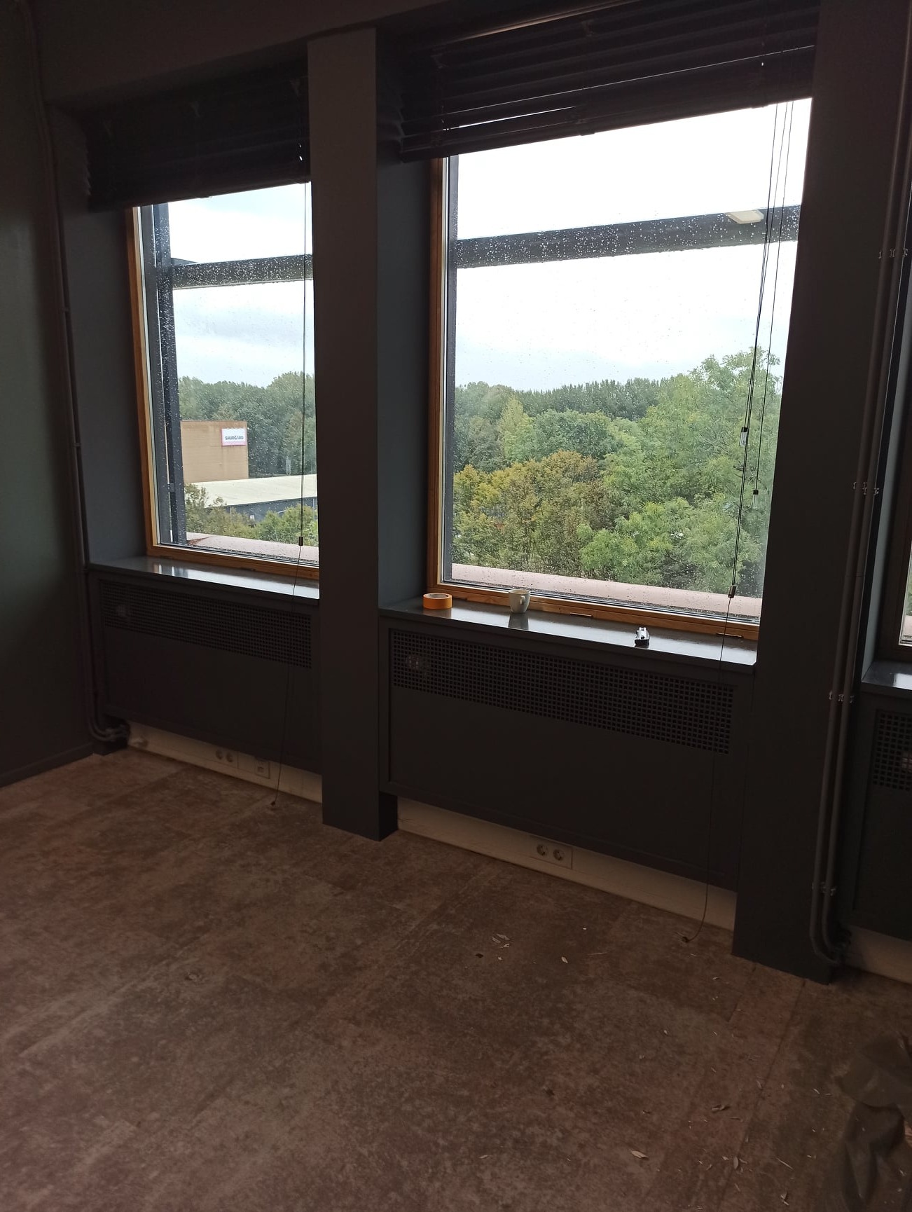 Pokój przed remontem: szare ściany, dwa okna z widokiem na zieleń za oknem, grzejniki pod oknami, widoczne gniazdka elektryczne, podłoga przygotowana do położenia nowej nawierzchni.