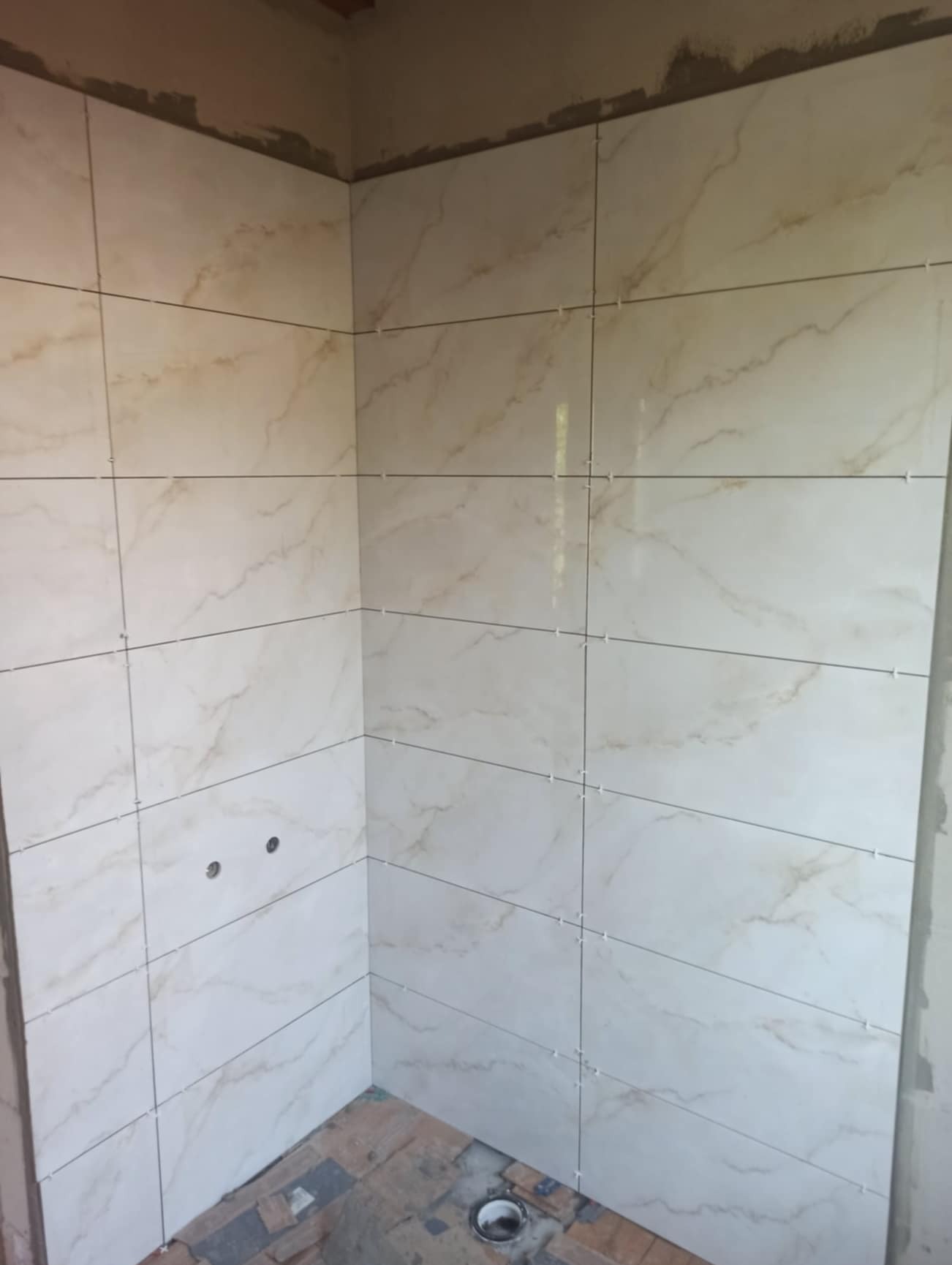 Ściana w rogu łazienki wyłożona jasnymi płytkami imitującymi marmur, z widocznymi krzyżykami dystansowymi i dwoma otworami na instalacje, w trakcie prac wykończeniowych.