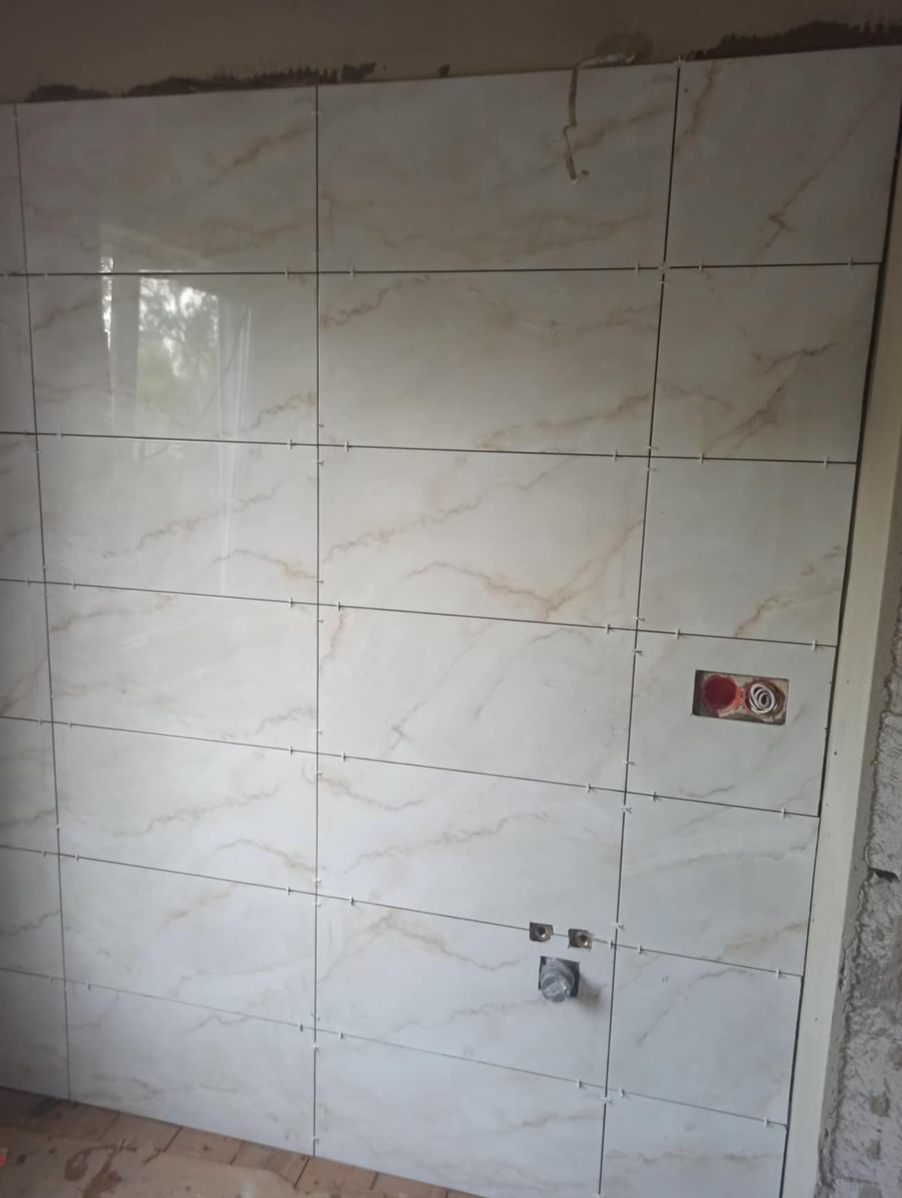 Ściana wyłożona jasnymi, marmurkowymi płytkami z krzyżykami dystansowymi, widoczne otwory na gniazdka elektryczne i rury wodne, surowy stan wykończenia.