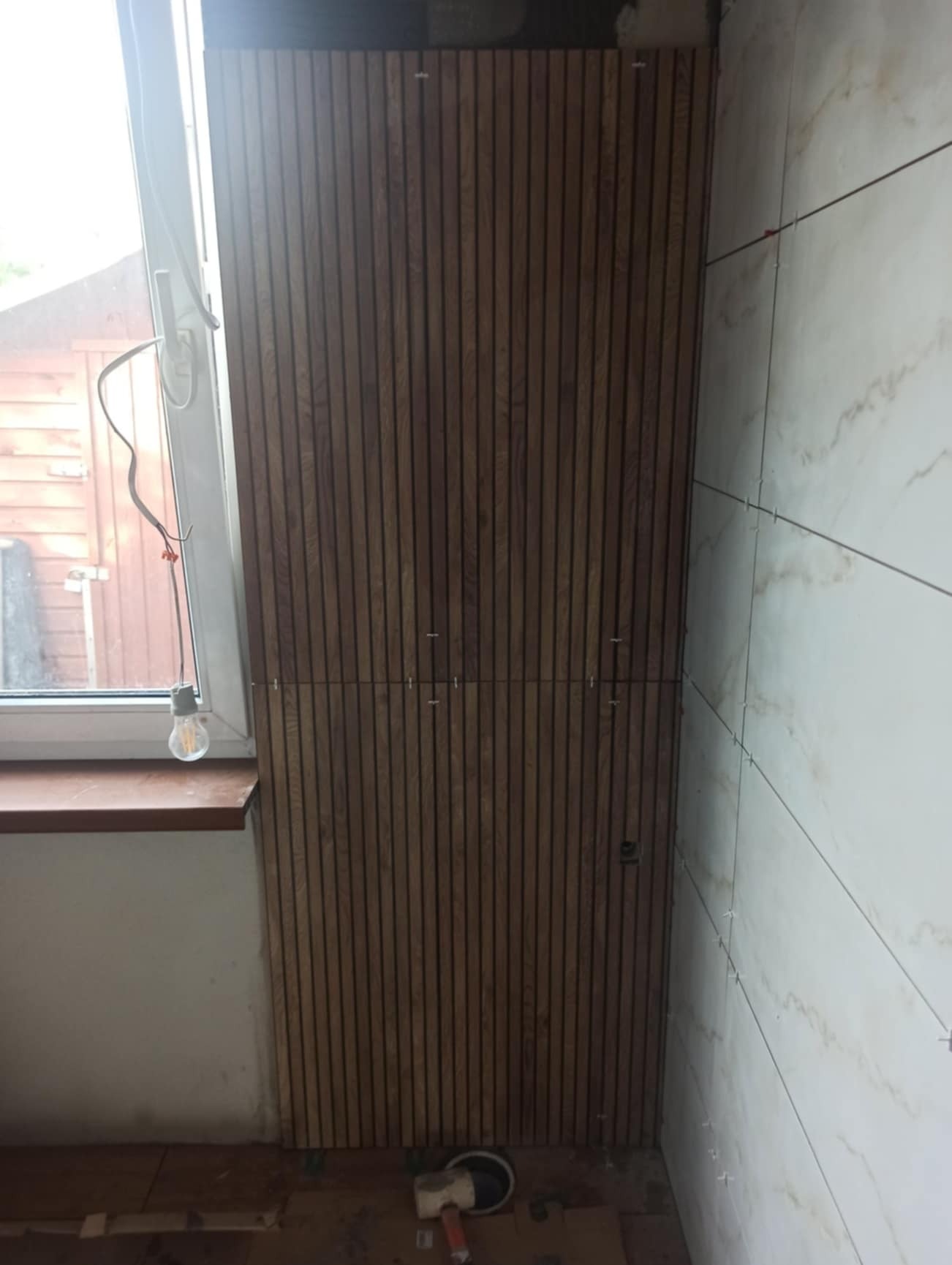 Fragment łazienki w trakcie prac: drewniane panele lamelowe na ścianie obok okna z żarówką, oraz płytki imitujące marmur na sąsiedniej ścianie, widoczne krzyżyki dystansowe.