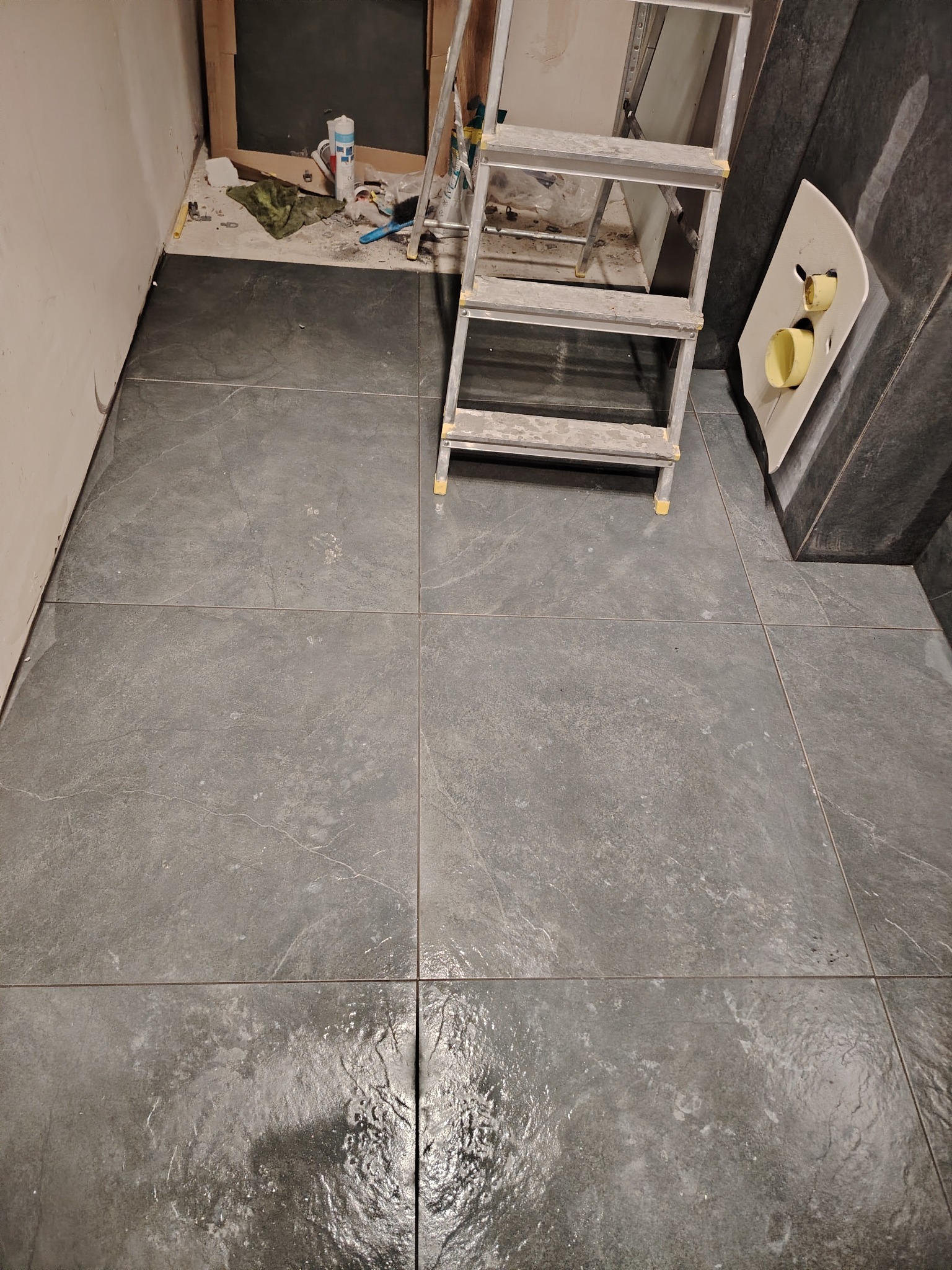 Ułożona szara podłoga z płytek w trakcie prac wykończeniowych w łazience, widoczna drabina, fragment instalacji sanitarnej i narzędzia pracy.