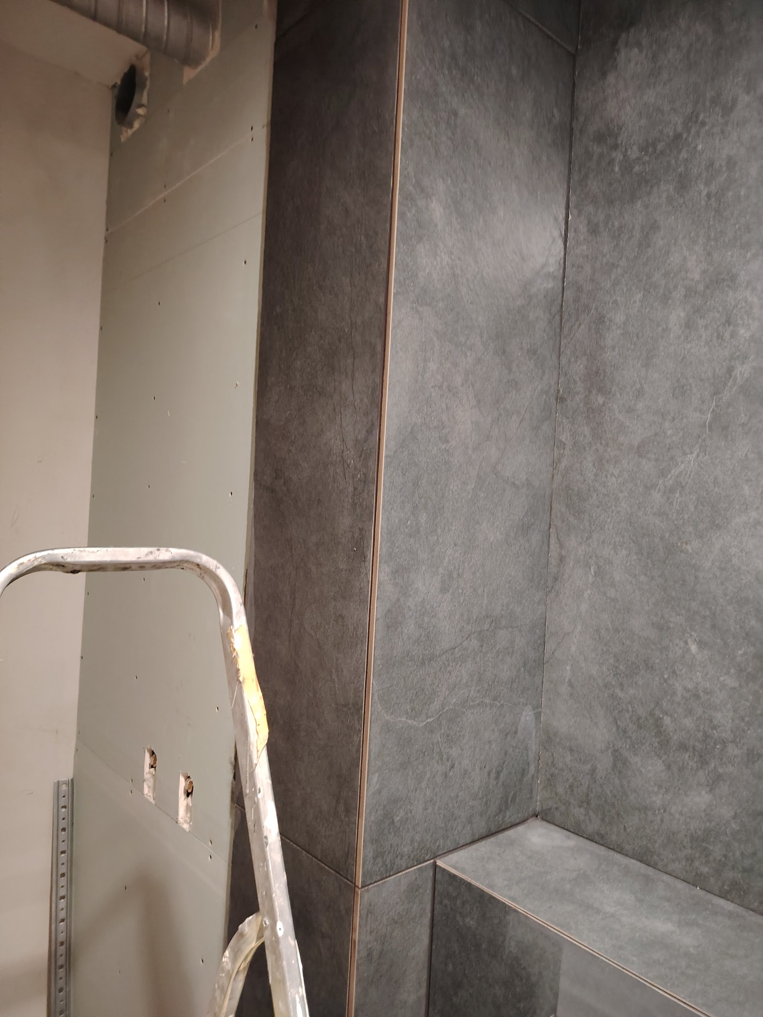 Wnętrze łazienki w trakcie prac wykończeniowych: ściana z płyt kartonowo-gipsowych z widocznymi otworami na gniazdka elektryczne, fragment drabiny malarskiej i narożnik wyłożony grafitowymi...