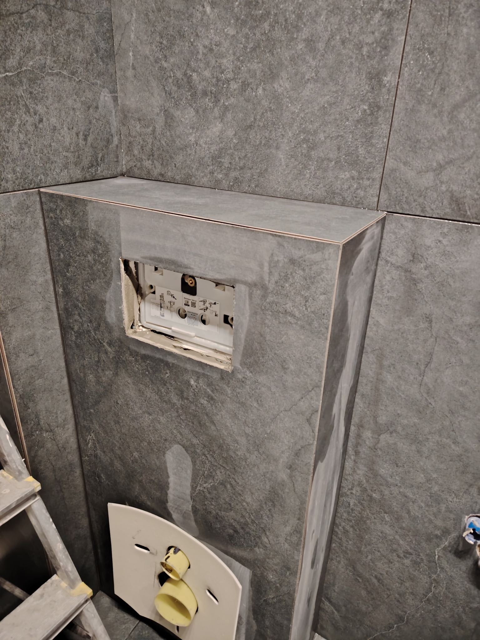 Montaż stelaża podtynkowego WC na tle szarych płytek imitujących kamień, widoczne fragmenty drabiny i elementów instalacji.