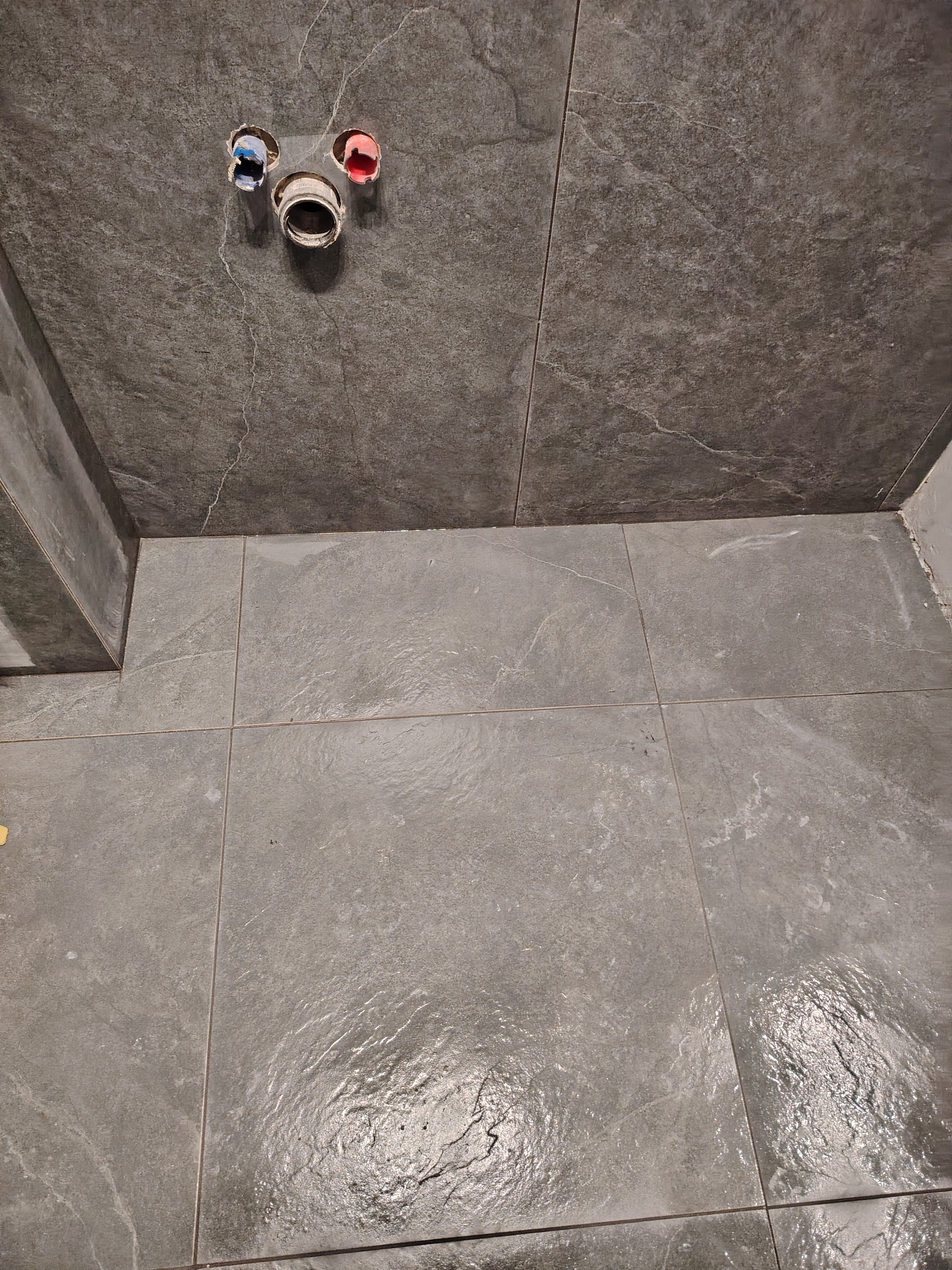 Szare płytki podłogowe i ścienne w łazience, widoczne przyłącza wodne z oznaczeniami kolorystycznymi, stan surowy przed montażem armatury.