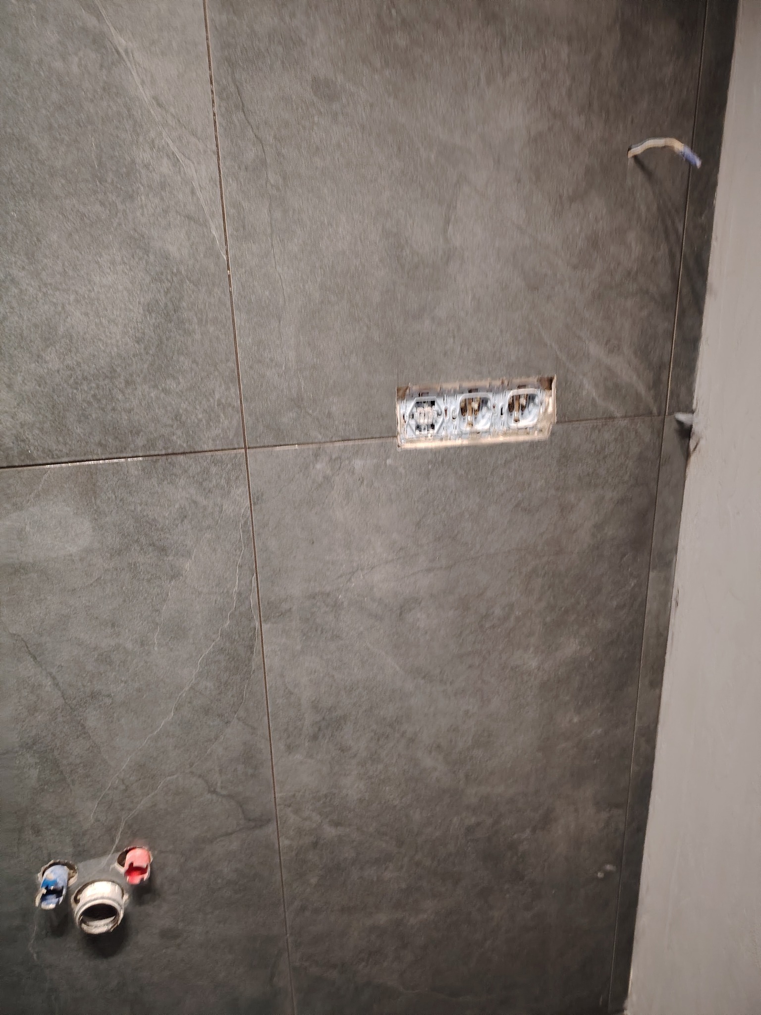 Szare płytki ceramiczne imitujące kamień na ścianie w łazience, widoczne otwory na gniazdka elektryczne i rury wodne przed montażem armatury.