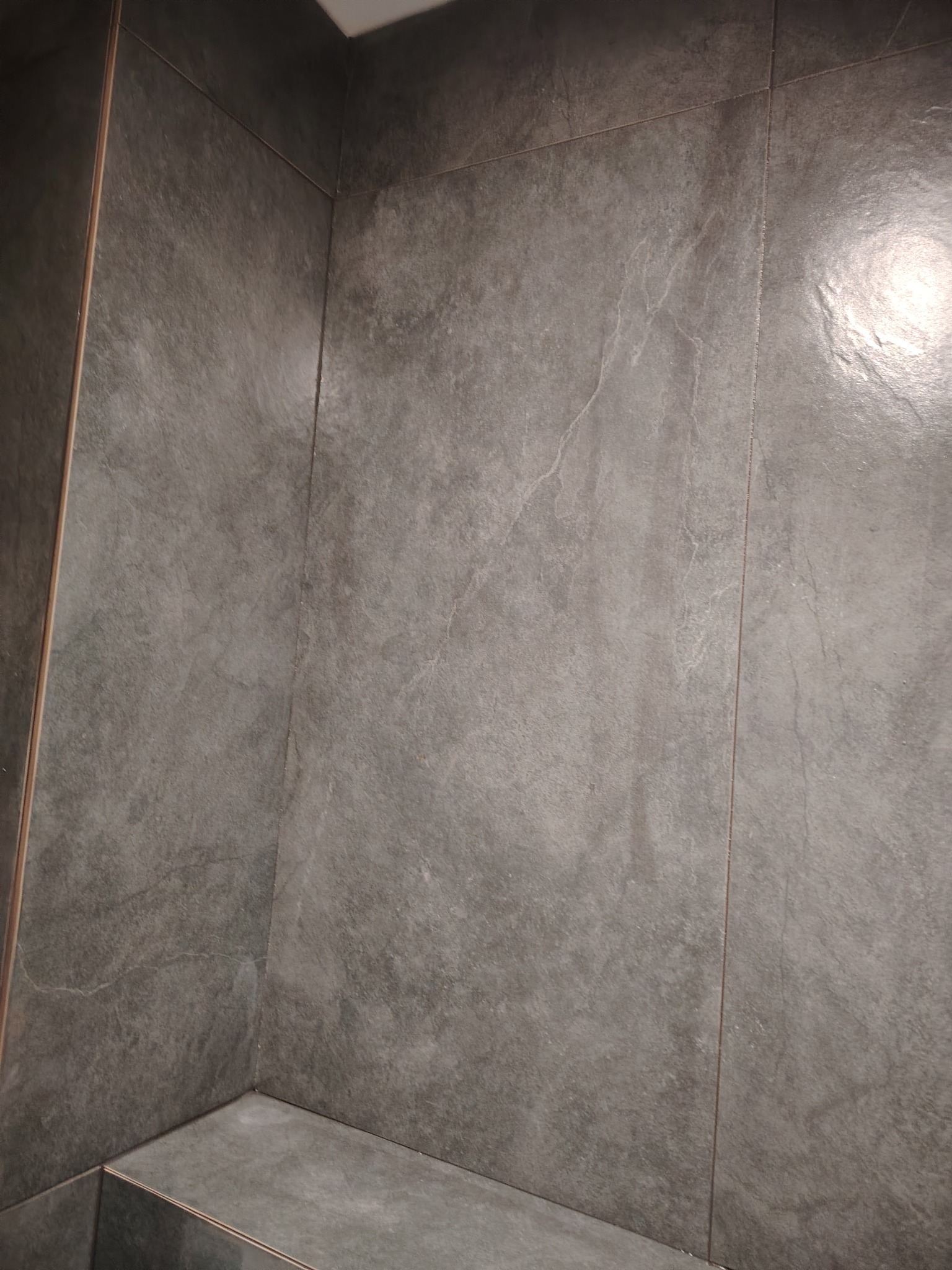 Fragment ściany wykończonej szarymi, matowymi płytkami imitującymi kamień, z widocznymi fugami i narożnikiem, oświetlonym górnym światłem.