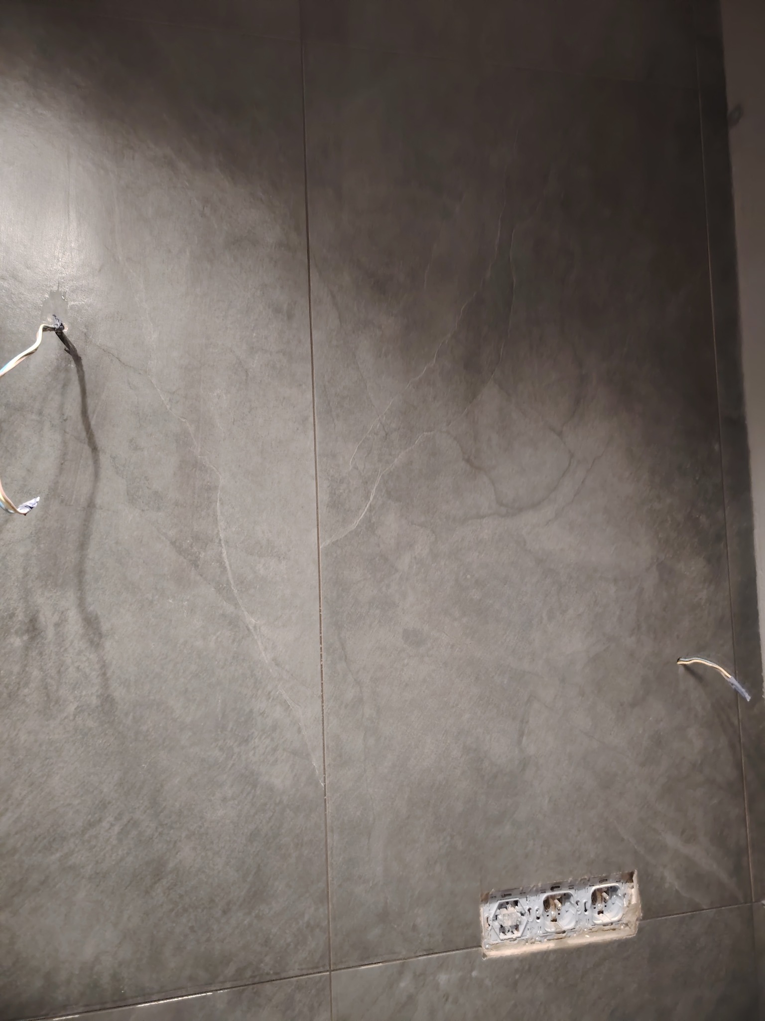 Ściana wyłożona szarymi płytkami imitującymi kamień, z widocznymi wystającymi przewodami i gniazdkami elektrycznymi bez obudowy, w trakcie prac wykończeniowych.