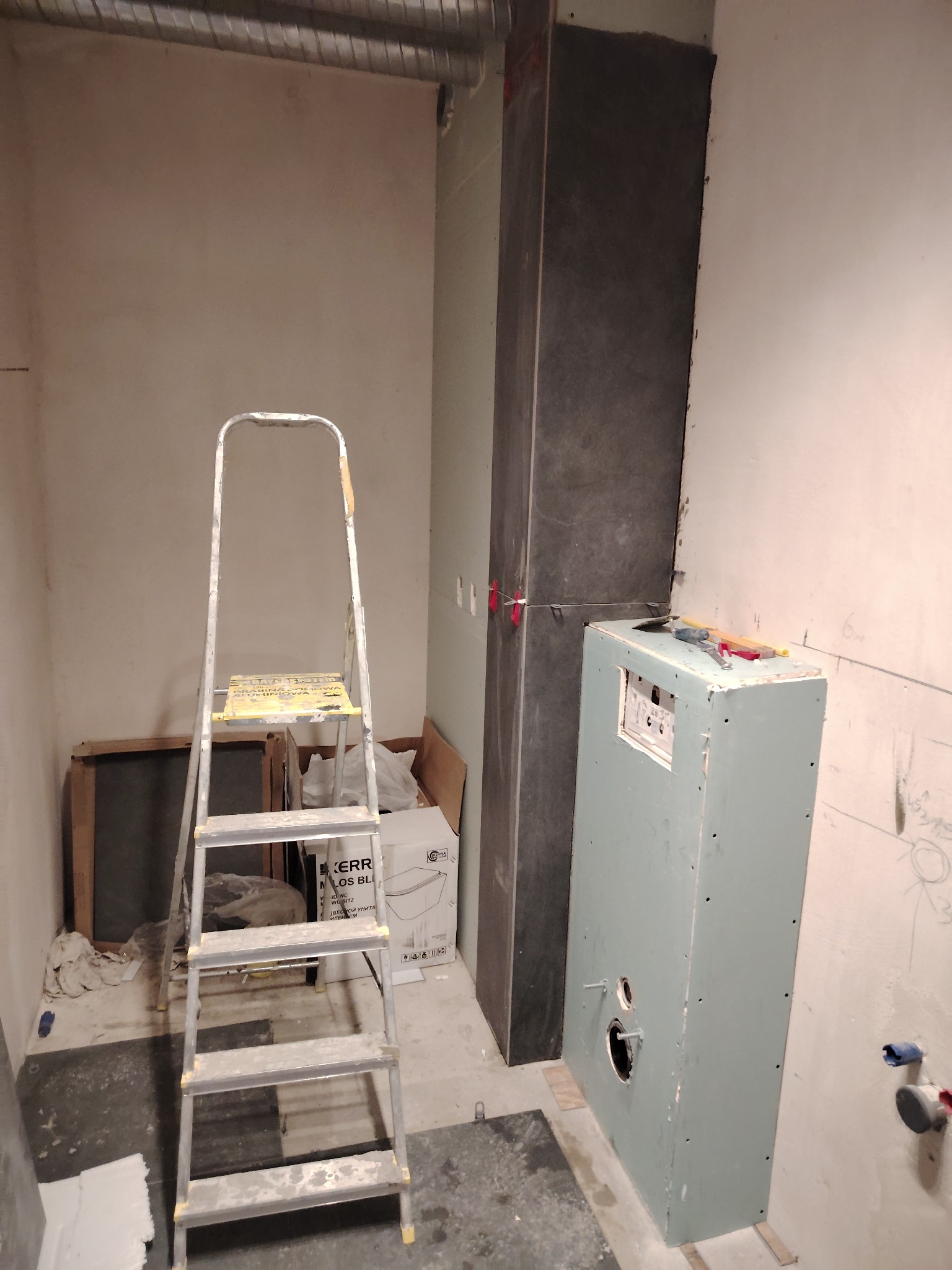 Wnętrze łazienki w trakcie prac wykończeniowych: widoczna drabina, obudowa stelaża WC z płyt karton-gips, fragment ściany z surową gładzią i instalacją hydrauliczną, oraz pion wentylacyjny obłożony...