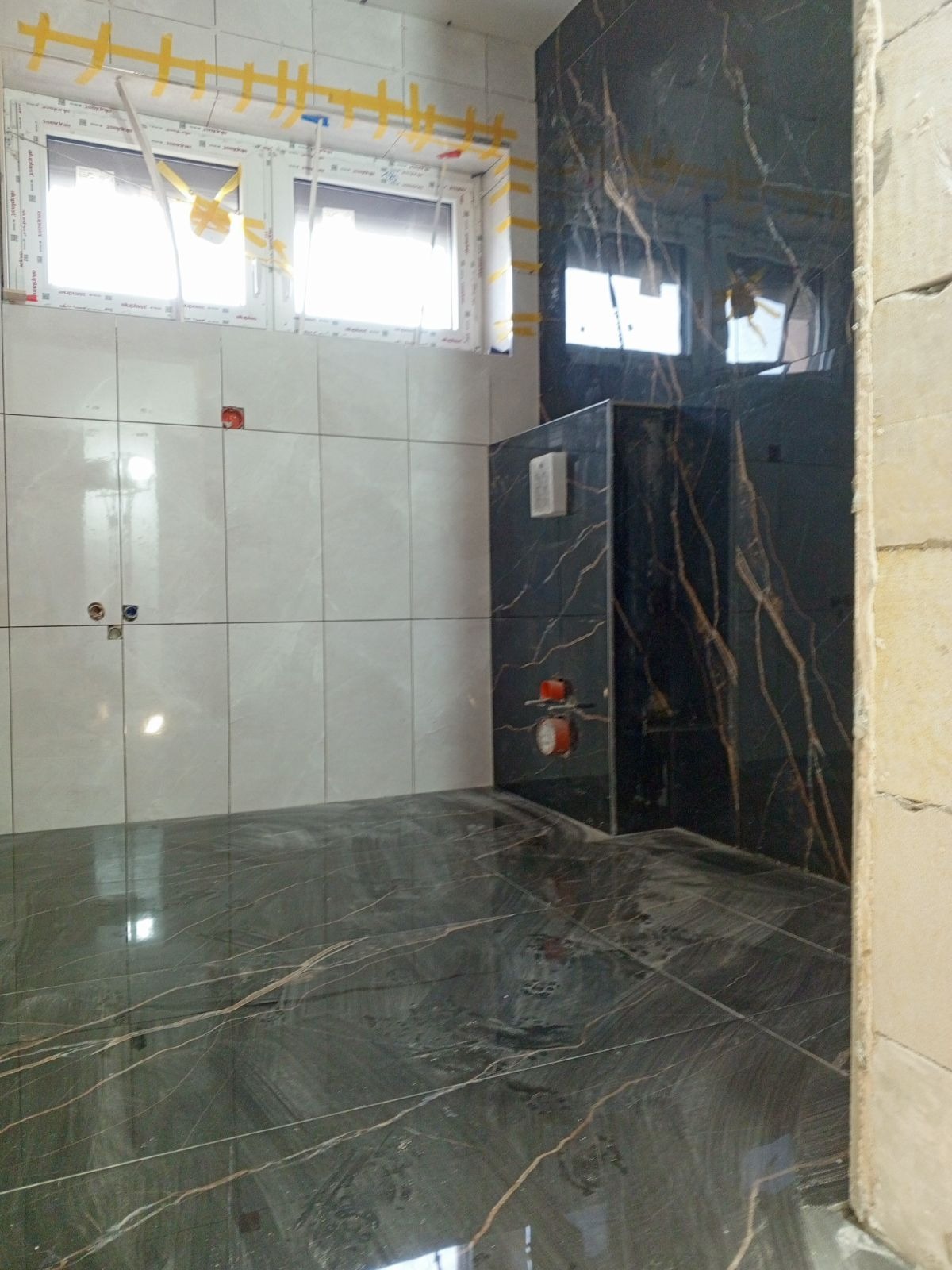 Niedokończona łazienka z czarnymi płytkami imitującymi marmur na podłodze i w strefie prysznica, białe płytki na ścianach, okno zabezpieczone taśmą malarską.