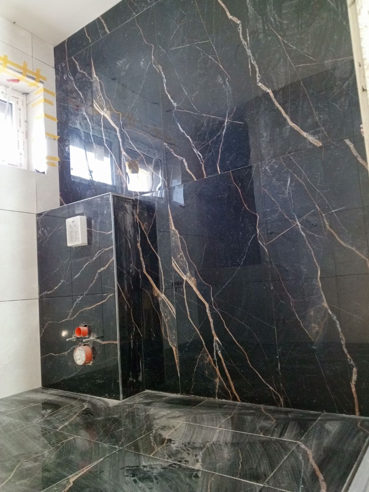Ciemne, marmurowe płytki z wyraźnym, złotym żyłowaniem na ścianie i podłodze łazienki w trakcie wykańczania, widoczne wystające rury instalacji wodnej i fragment okna z taśmą malarską.