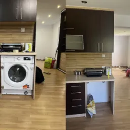 Kuchenna zabudowa z brązowymi szafkami i pralką w zabudowie, widok na otwartą przestrzeń z podłogą panelową i elementami wyposażenia w tle.