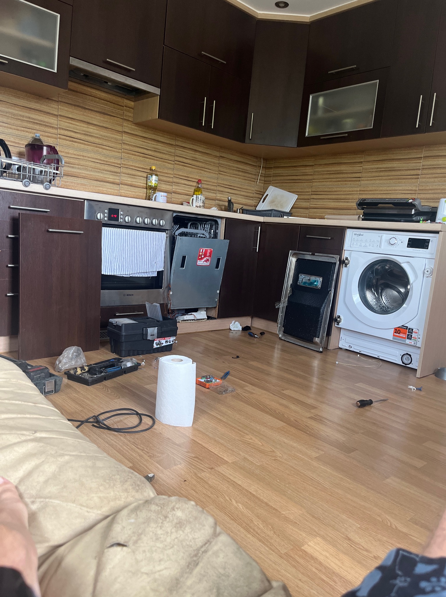 Kuchnia w trakcie naprawy: otwarty zmywarka i pralka, skrzynka z narzędziami, rozłożone narzędzia na podłodze z panelami, widoczne elementy zabudowy kuchennej.