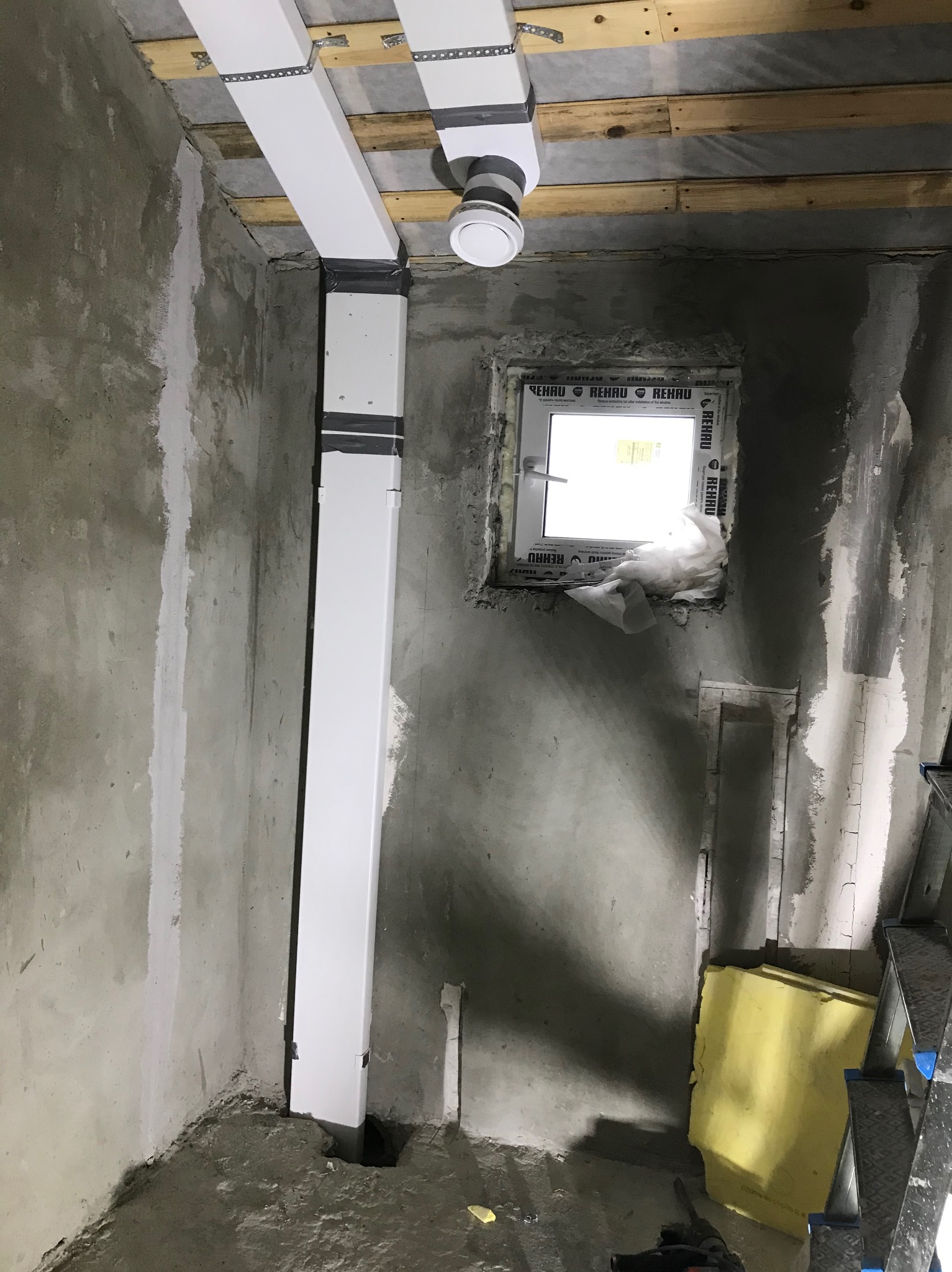 Wnętrze w trakcie remontu: widoczne surowe ściany, okno w stanie montażu z widoczną pianką uszczelniającą, białe rury wentylacyjne pod sufitem, częściowo zaklejone taśmą oraz fragment drabiny...