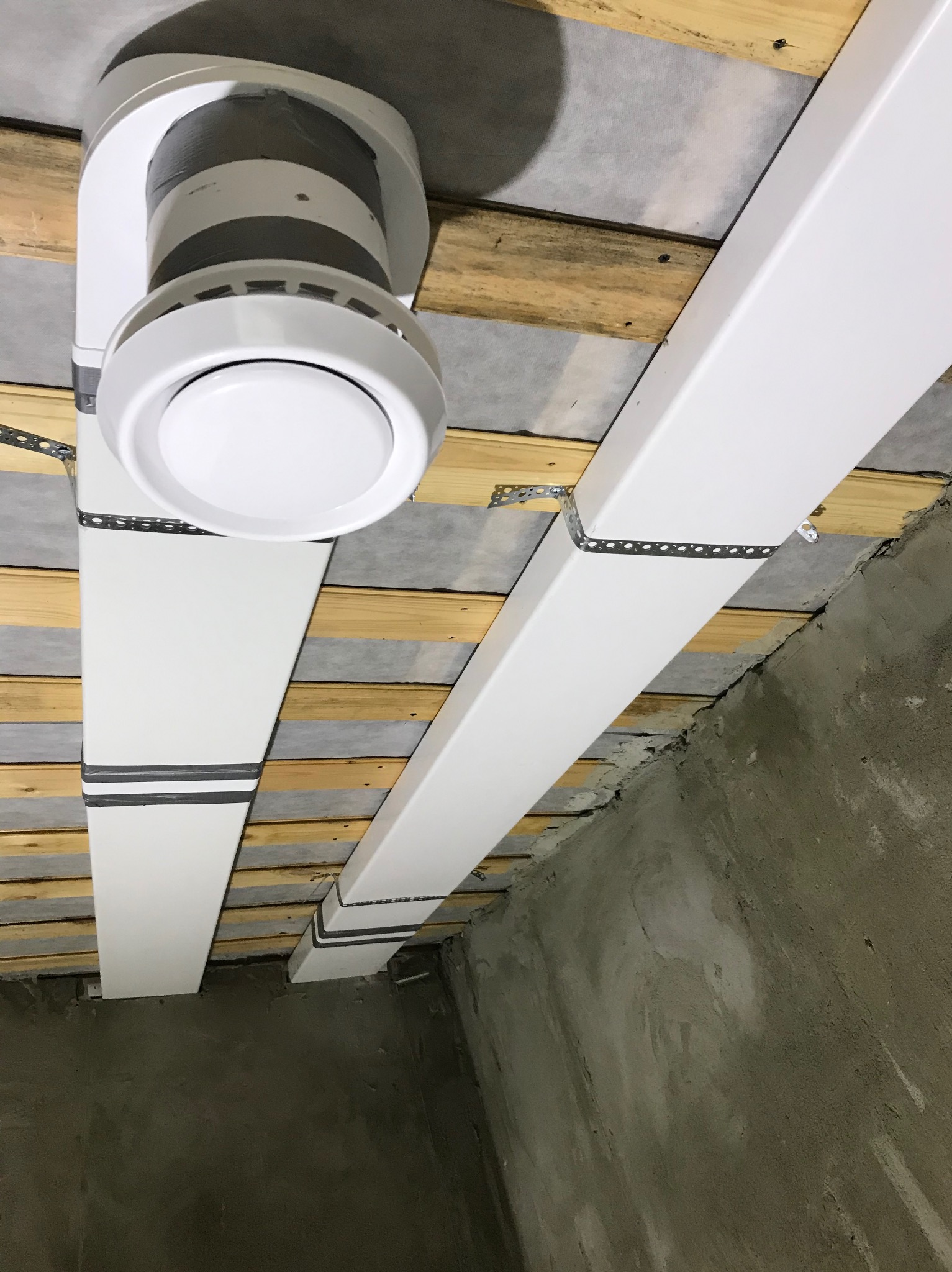 Instalacja sufitowa z białymi belkami i okrągłym elementem wentylacyjnym, widok z dołu na tle surowego betonu.