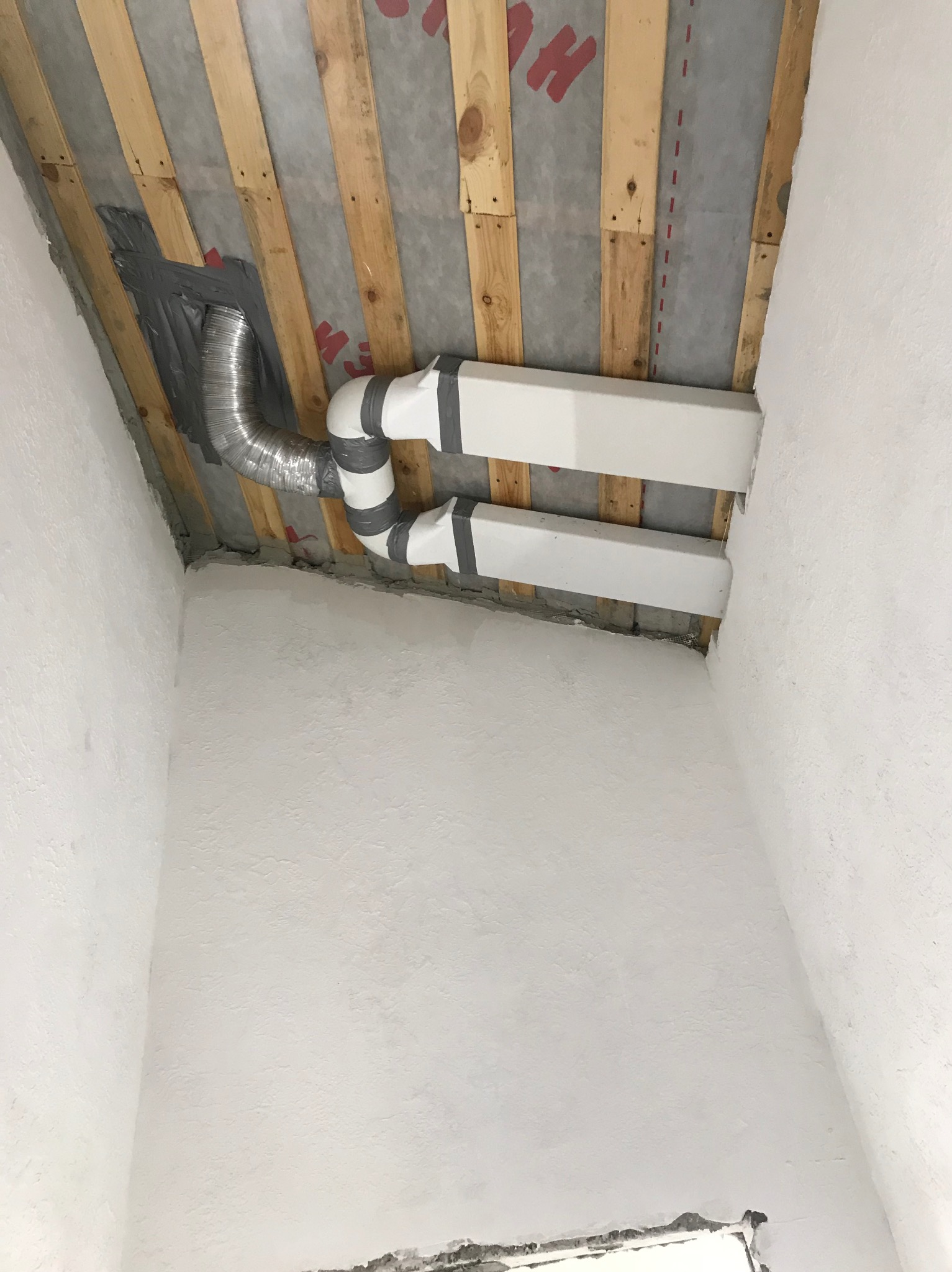 Fragment wnętrza w trakcie remontu: białe ściany z surowym wykończeniem, widoczne drewniane belki stropowe z izolacją, fragment instalacji wentylacyjnej z rurą aluminiową i białymi kanałami, całość...