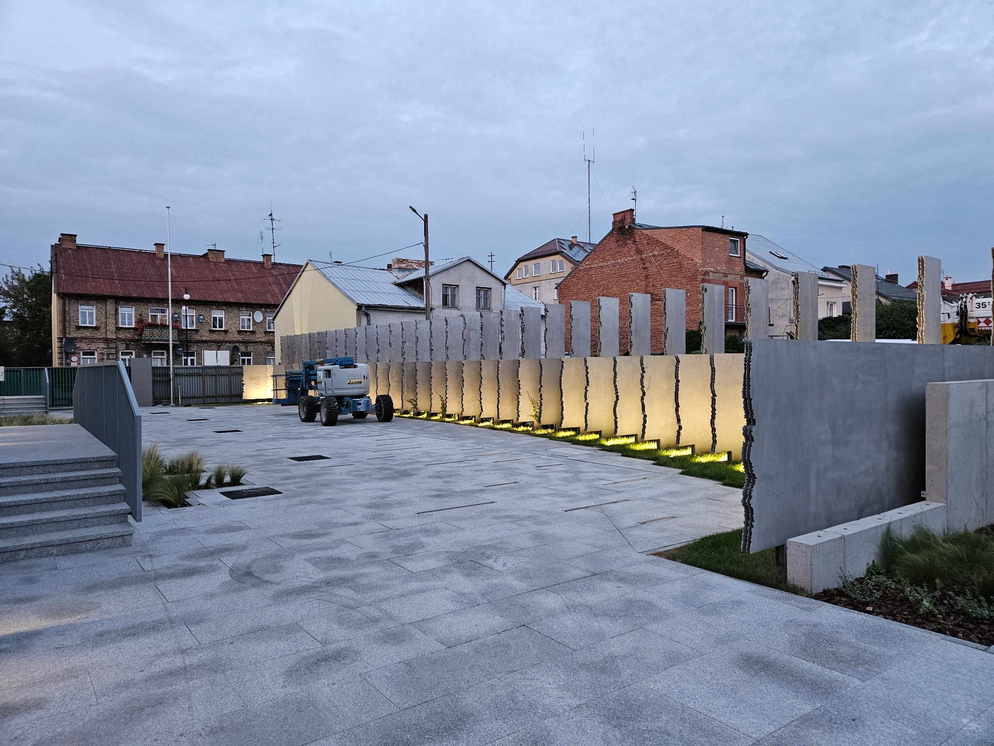 Oświetlony plac z betonowymi elementami dekoracyjnymi i podnośnikiem koszowym w tle, widok na budynki mieszkalne.