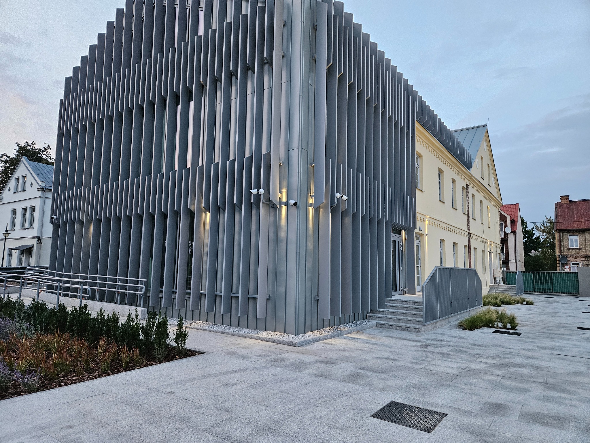 Nowoczesny budynek z pionowymi panelami elewacyjnymi i systemem kamer monitoringu, widok z zewnątrz na tle starszej zabudowy.