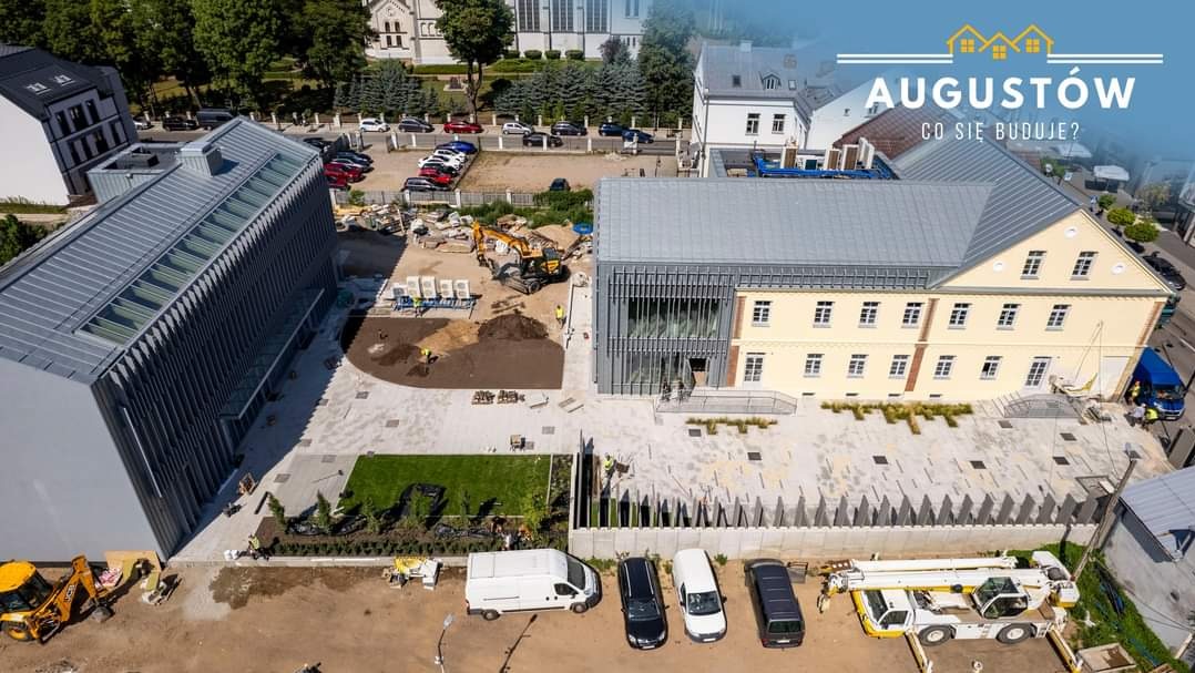 Widok z góry na plac budowy w Augustowie z dwoma budynkami o nowoczesnej architekturze, żółtą koparką i zaparkowanymi samochodami dostawczymi oraz dźwigiem.