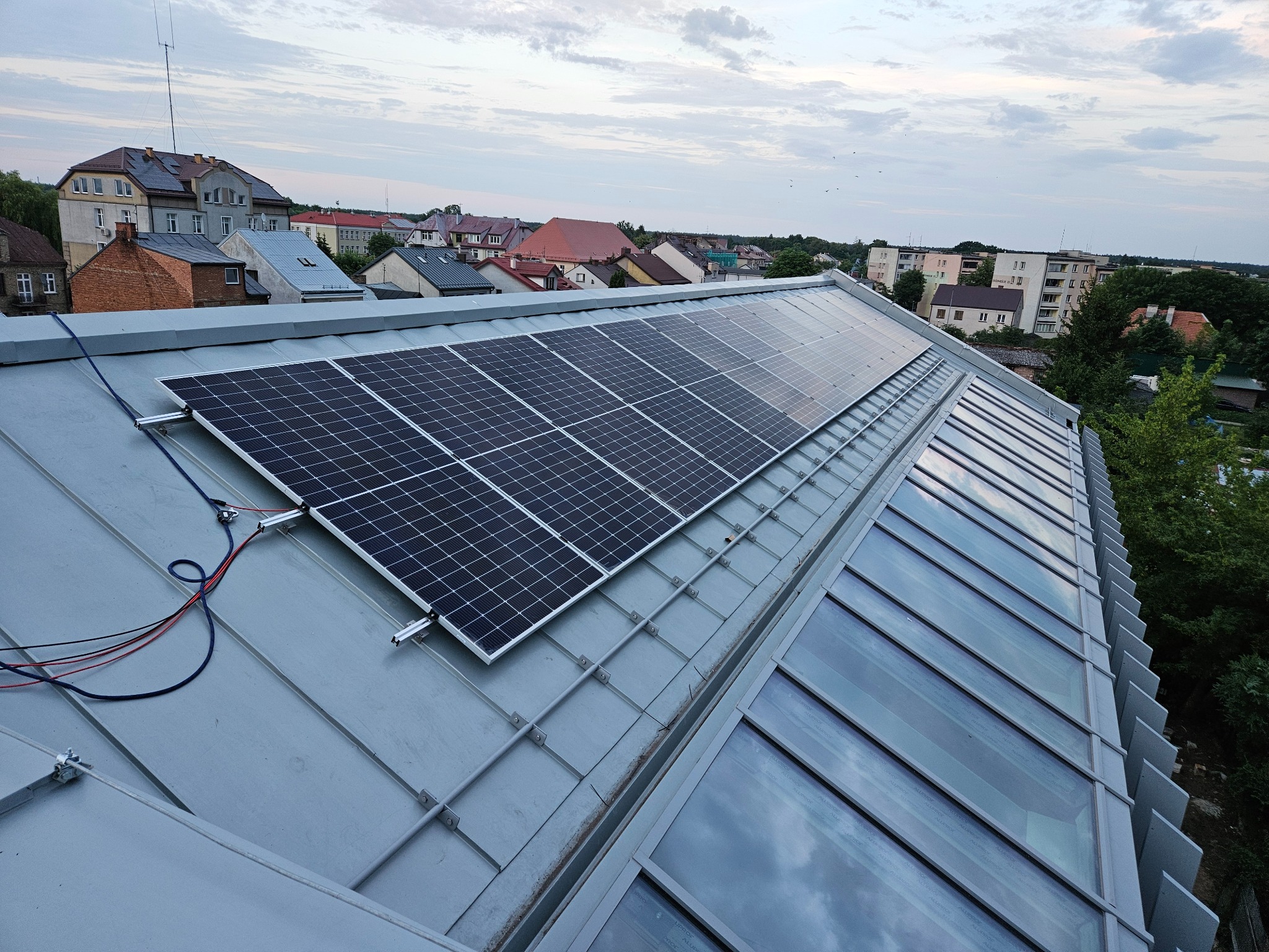 Panel słoneczny zamontowany na metalowym dachu budynku mieszkalnego, widok z góry na tle zabudowy miejskiej i pochmurnego nieba.