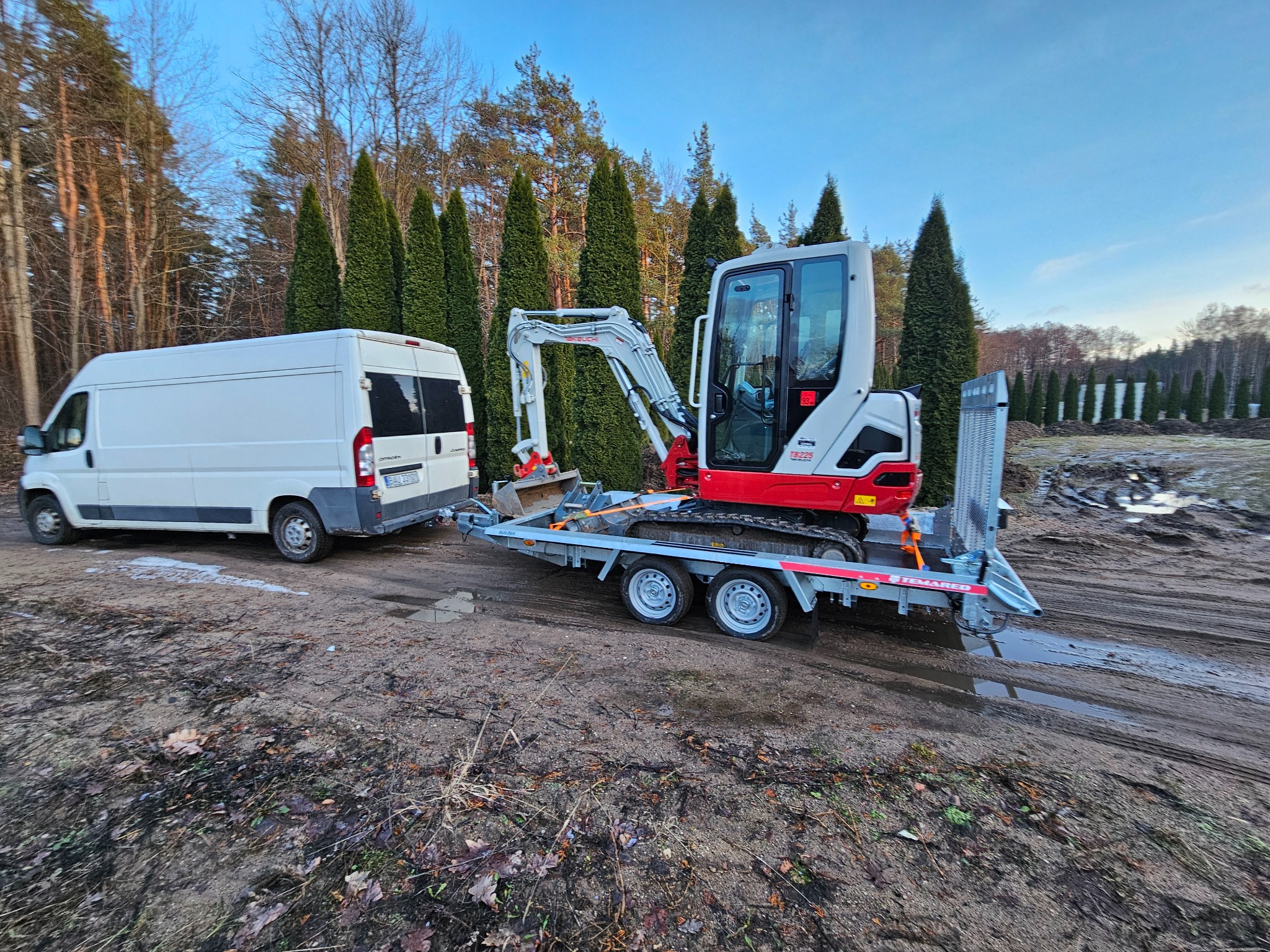 Biały bus z przyczepą przewożący mini koparkę na tle drzew iglastych i błotnistego terenu.