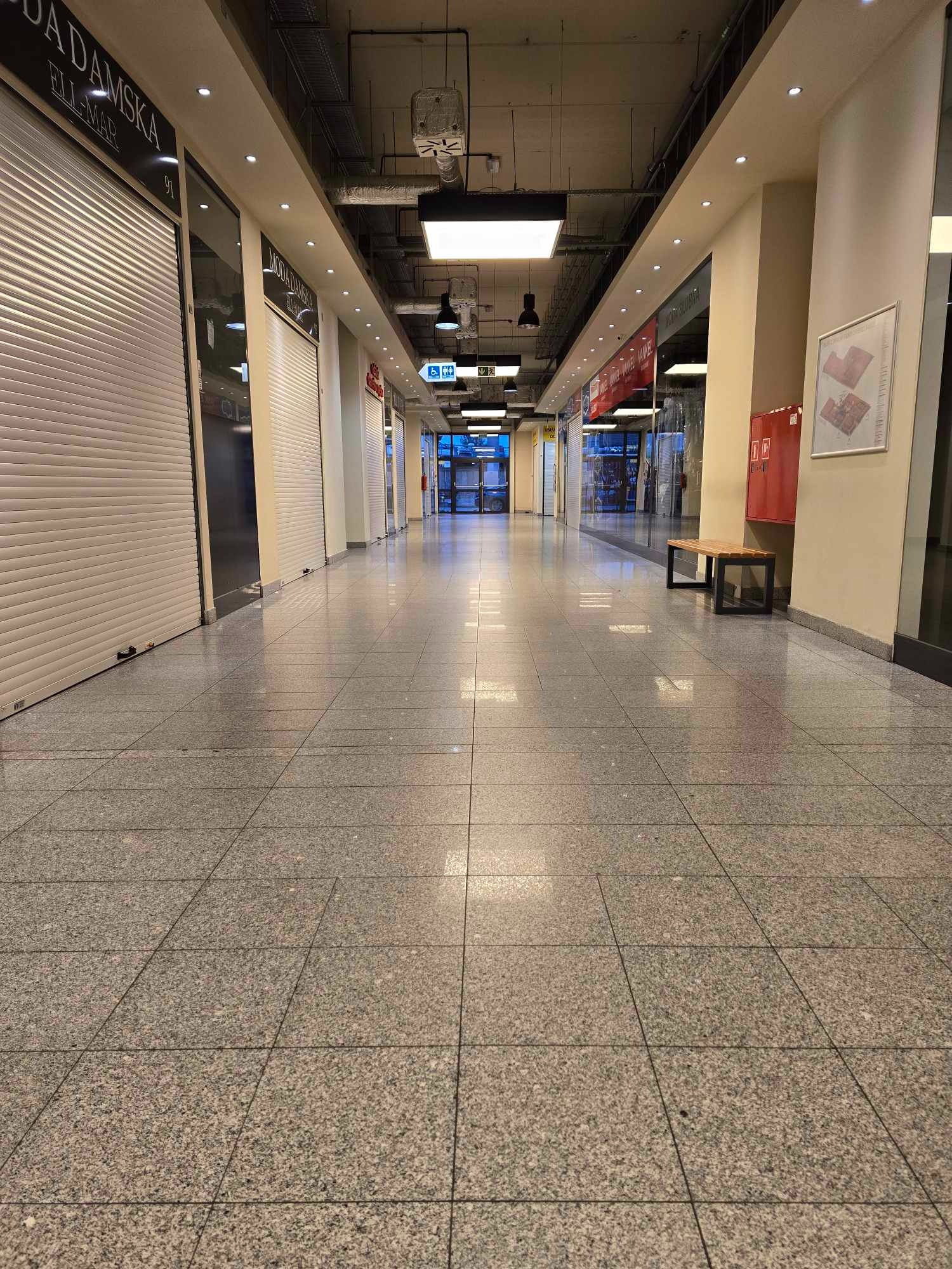 Pusta galeria handlowa z zamkniętymi sklepami, widok na korytarz z szarymi płytkami podłogowymi i roletami na sklepach, oświetlenie sufitowe.