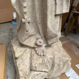 Detal rzeźby sakralnej z piaskowca: fałdy szat, różaniec z krzyżem, stopa z różą, surowa faktura kamienia. Widoczne ślady dłuta, warsztatowy charakter ujęcia.