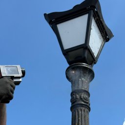 Pure Laser - Piaskowanie zabytkowej latarni ulicznej. W tle błękitne niebo. Operator trzyma w dłoni pistolet do piaskowania. Widoczne ślady czyszczenia.