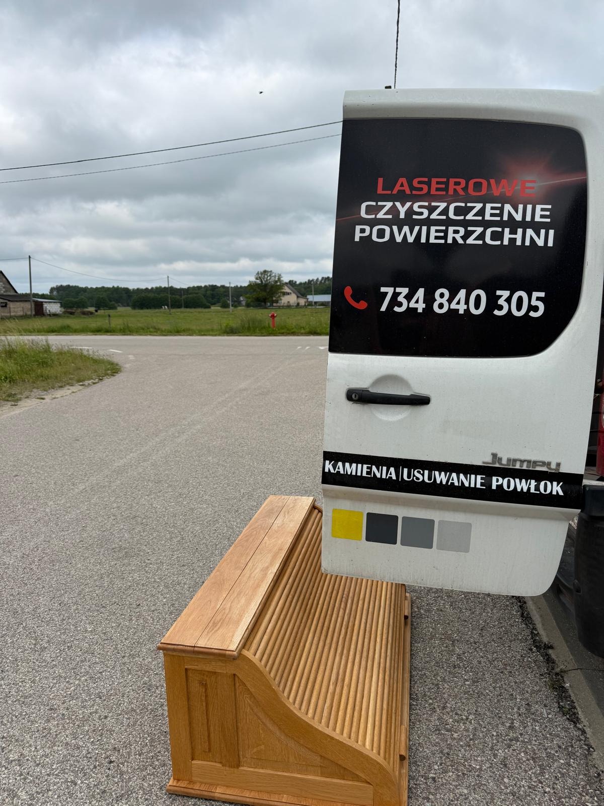 Otwarta tylna klapa busa z reklamą laserowego czyszczenia powierzchni i numerem telefonu, obok drewniana szafka ze żłobieniami, na tle drogi i zabudowań.