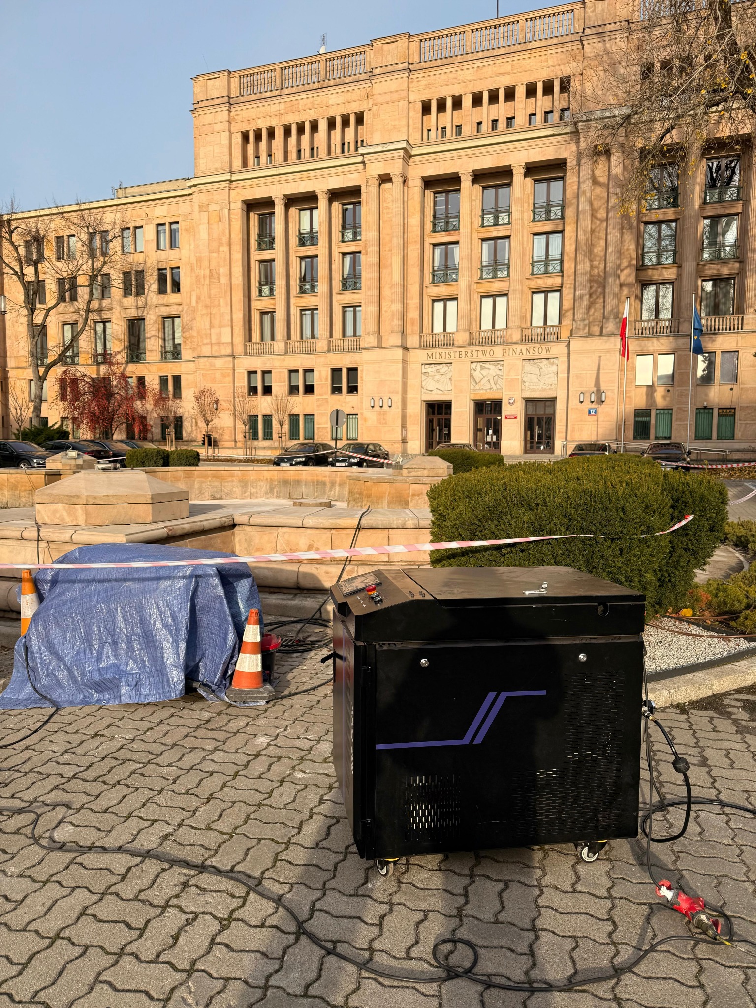 Czarna maszyna czyszcząca na kostce brukowej, z budynkiem Ministerstwa Finansów w tle. Fontanna zabezpieczona taśmą i plandeką. Widoczne kable i stożki.