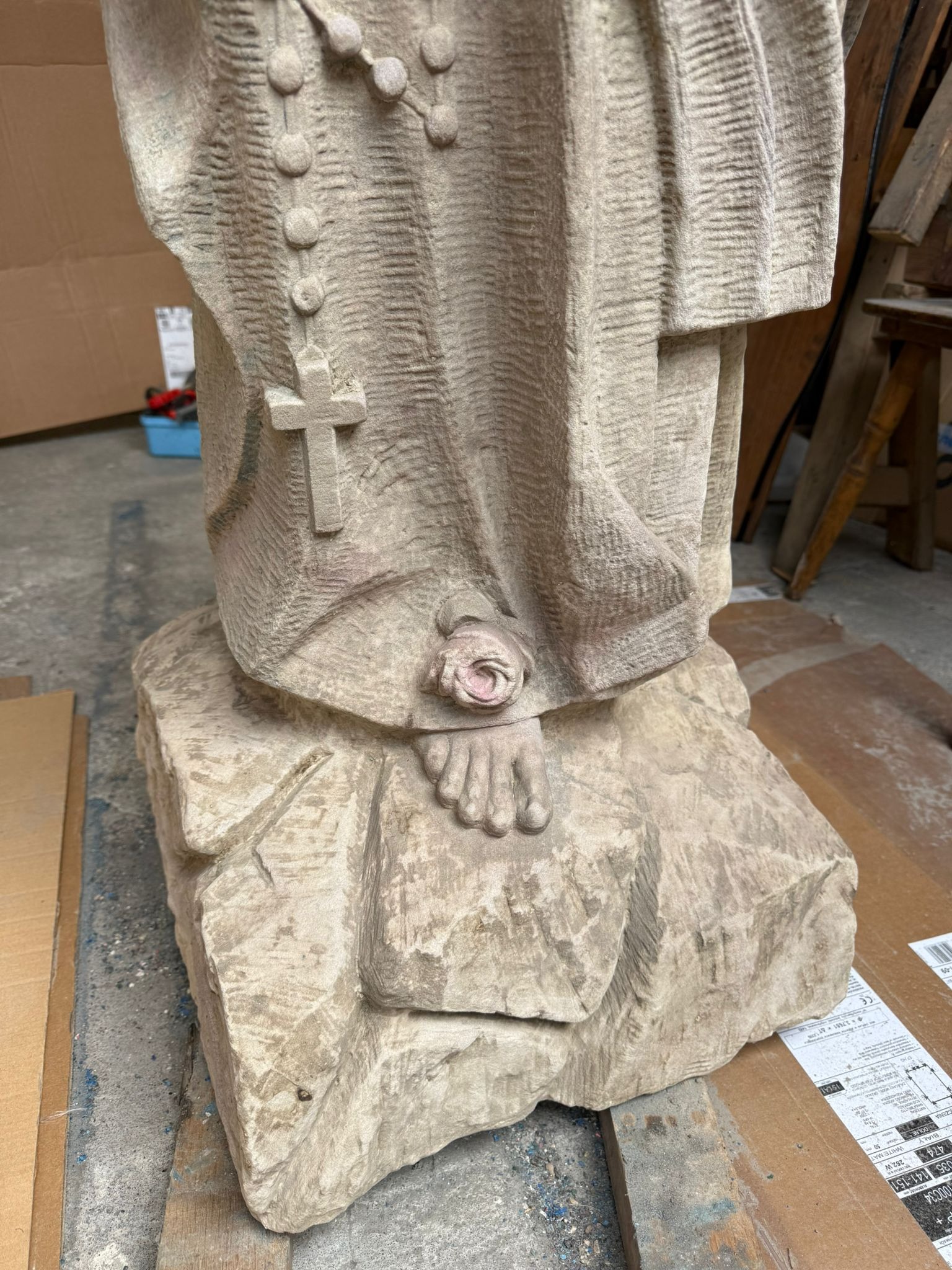 Detal rzeźby sakralnej z piaskowca: fałdy szat, różaniec z krzyżem, stopa z różą, surowa faktura kamienia. Widoczne ślady dłuta, warsztatowy charakter ujęcia.