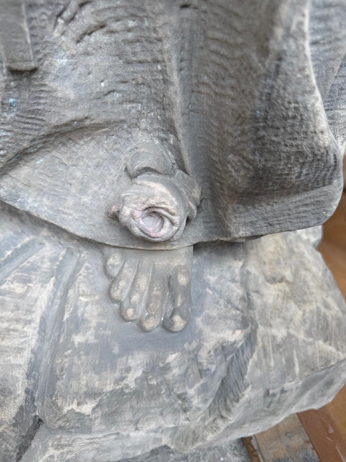 Fragment kamiennej rzeźby: stopa i dłoń postaci w szatach, widoczne detale obróbki kamienia i specyficzna faktura materiału.