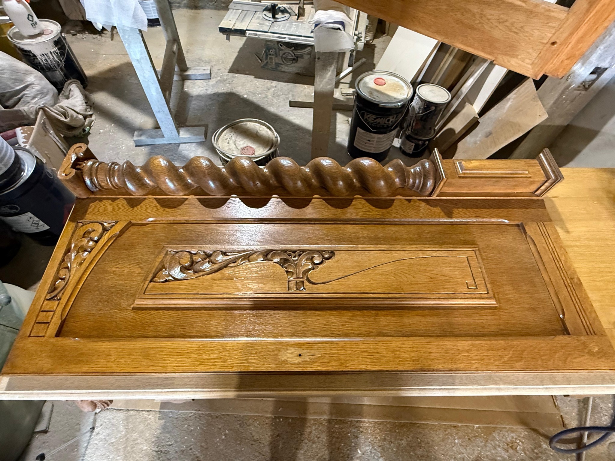 Odrestaurowany, drewniany element dekoracyjny z rzeźbieniami, leżący na stole warsztatowym. Widoczne puszki z farbą i narzędzia w tle. Ciepłe oświetlenie podkreśla detale.