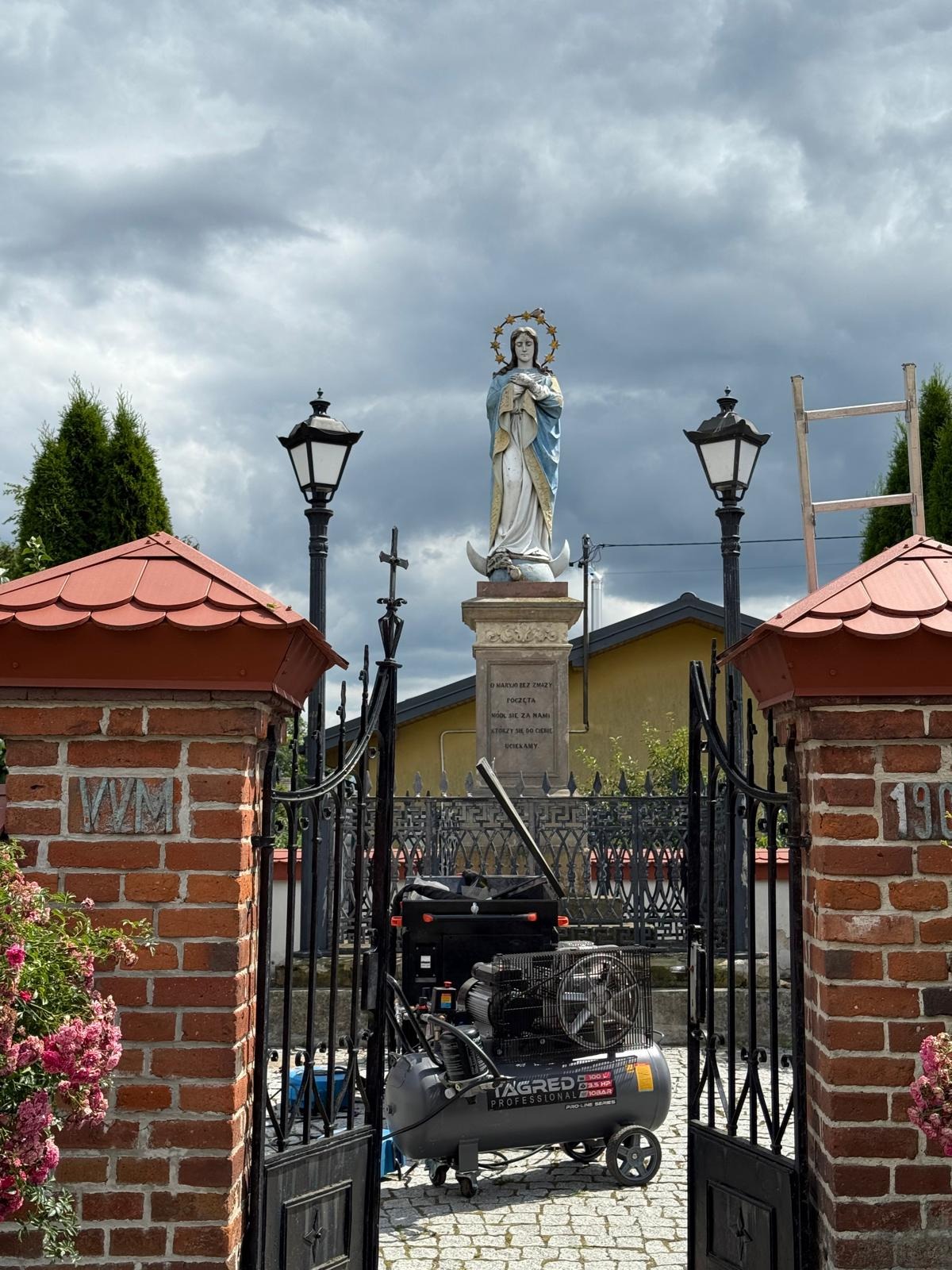 Statua Matki Boskiej z kompresorem u stóp, widok zza otwartej, ozdobnej bramy. W tle pochmurne niebo i latarnie. Sprzęt sugeruje prace konserwatorskie.