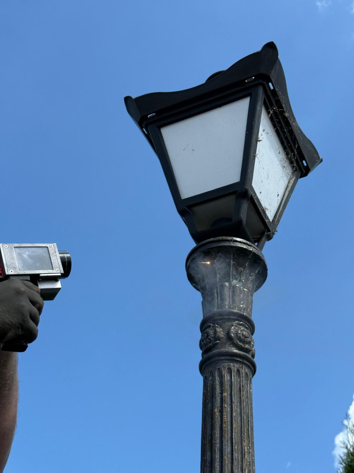 Piaskowanie zabytkowej latarni ulicznej. W tle błękitne niebo. Operator trzyma w dłoni pistolet do piaskowania. Widoczne ślady czyszczenia.