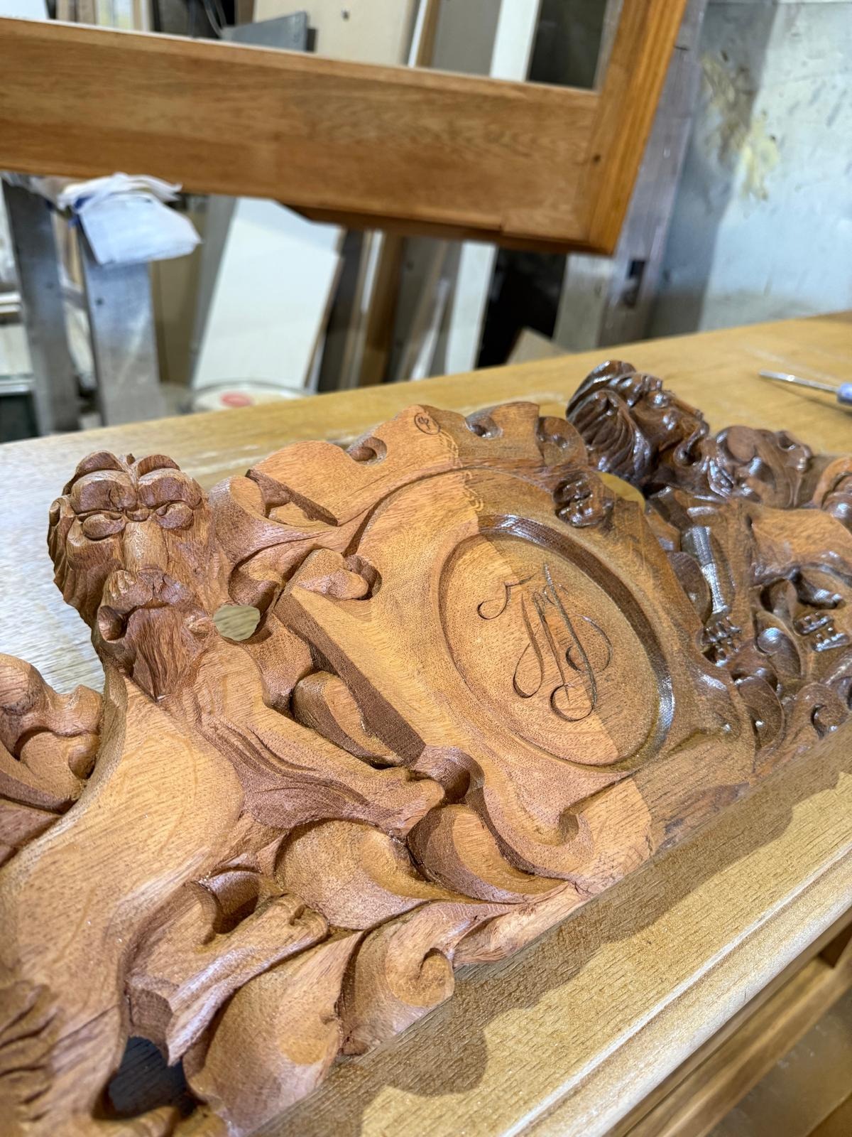 Drewniana płaskorzeźba z herbem i motywami roślinnymi, leżąca na drewnianym stole. Delikatne światło podkreśla detale rzeźby. Widoczne narzędzia w tle.