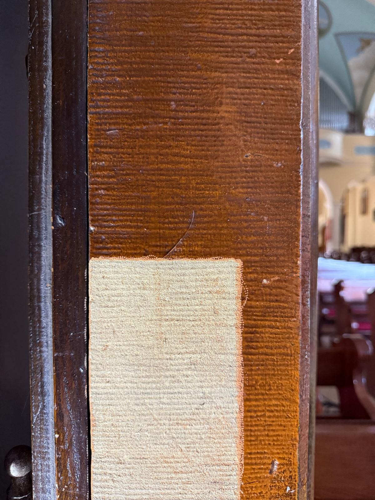 Fragment odnowionej, drewnianej ramy drzwi w kościele. Widoczny jaśniejszy, naprawiony obszar i oryginalna, brązowa faktura drewna. Detal konserwatorski.