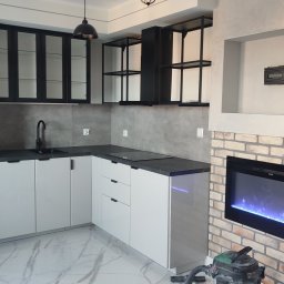Ruslan Iva - Nowoczesna, jasna kuchnia w Tczewie z białymi szafkami, czarnymi blatami i przeszklonymi szafkami wiszącymi, uzupełniona o dekoracyjny kominek elektryczny z ceglaną obudową.