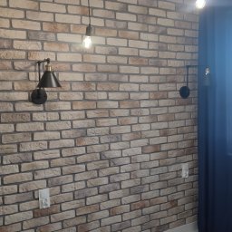 Ruslan Iva - Ściana z cegły dekoracyjnej z oświetleniem: czarne kinkiety i wiszące żarówki na kablach, marmurowa podłoga, fragment zasłony.