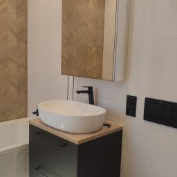 Ruslan Iva - Nowoczesna łazienka z białą umywalką nablatową na czarnej szafce, lustrem z drewnianym wzorem jodełki i podświetleniem LED pod szafką, widoczna toaleta i płytki z delikatnym wzorem.