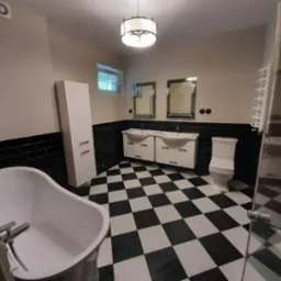 Czarno-biała łazienka w stylu retro z wanną wolnostojącą, dwiema umywalkami z szafkami, toaletą i czarnymi płytkami na ścianach, oświetlona lampą sufitową.