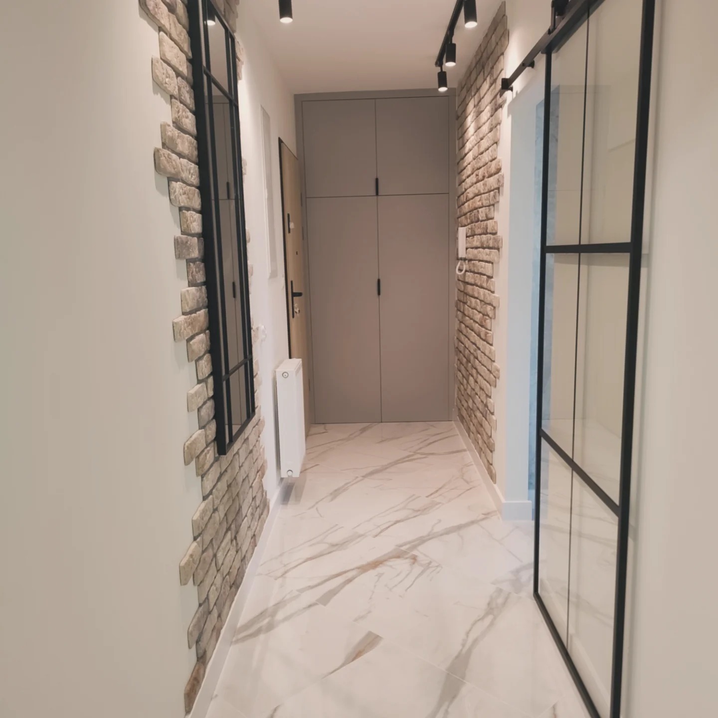 Korytarz z dekoracyjną ścianą z cegły, marmurową podłogą, przesuwnymi drzwiami z czarną ramą i szarą szafą wnękową.
