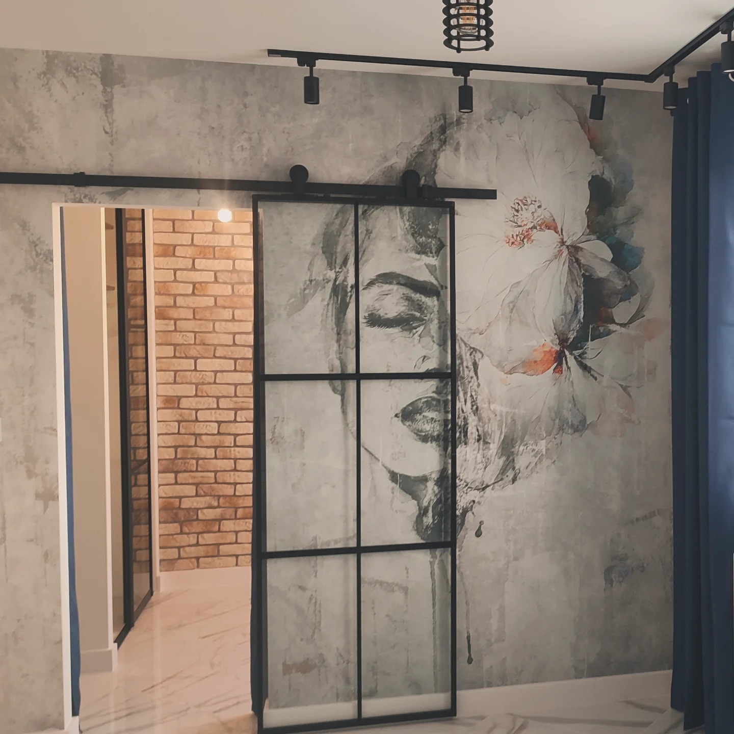 Przesuwne drzwi w stylu loft z czarną ramą, z motywem abstrakcyjnej twarzy i kwiatu na szarej ścianie z efektem betonu, oświetlenie szynowe na suficie, widoczny fragment ściany z cegły w tle.