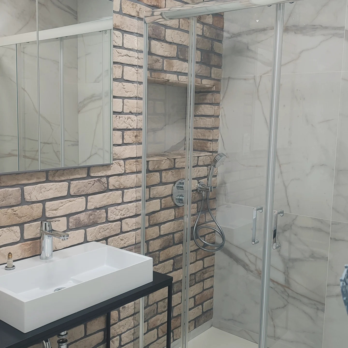 Nowoczesna łazienka z kabiną prysznicową ze szklanymi drzwiami, umywalką nablatową na czarnym stelażu i ścianą wyłożoną cegłą oraz płytkami imitującymi marmur.