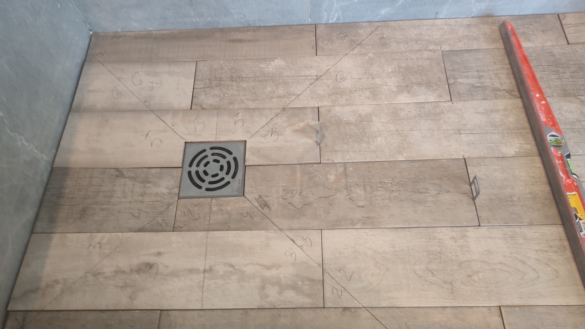 Podłoga łazienki wyłożona płytkami imitującymi drewno, z centralnie umieszczonym odpływem liniowym i zaznaczonymi liniami cięcia oraz numeracją, poziomica przy krawędzi.