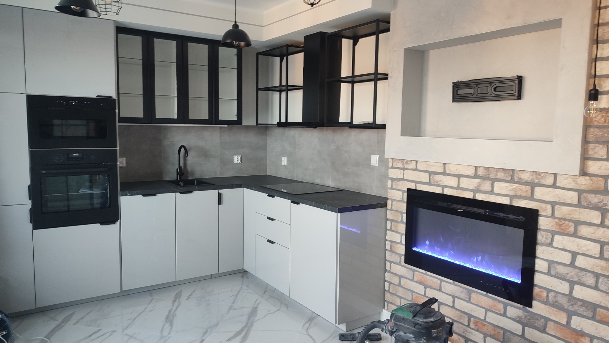 Nowoczesna, jasna kuchnia w Tczewie z białymi szafkami, czarnymi blatami i przeszklonymi szafkami wiszącymi, uzupełniona o dekoracyjny kominek elektryczny z ceglaną obudową.