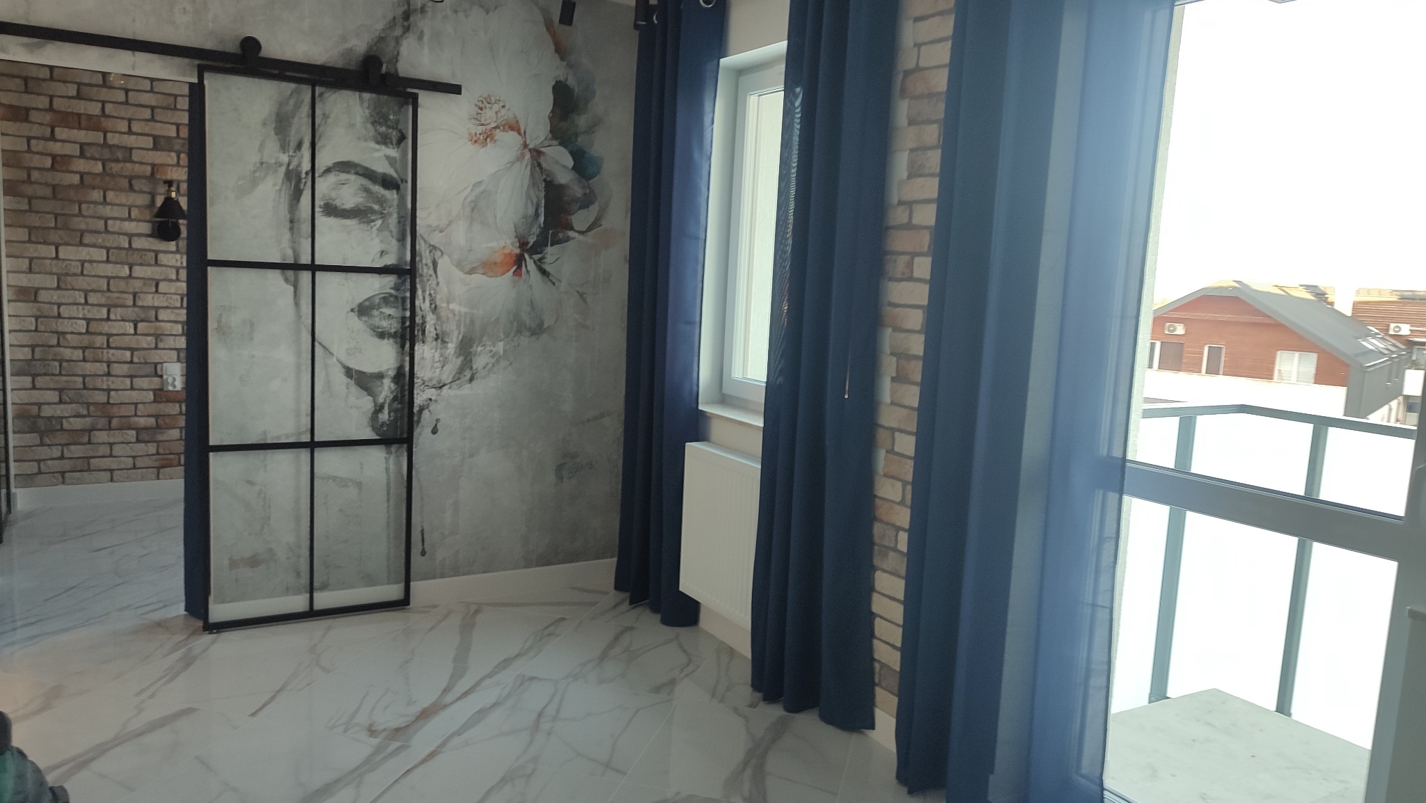 Nowoczesne wnętrze z przesuwnymi drzwiami loftowymi z grafiką, marmurową podłogą, ścianą z cegły i zasłonami w odcieniach granatu, widok na balkon.