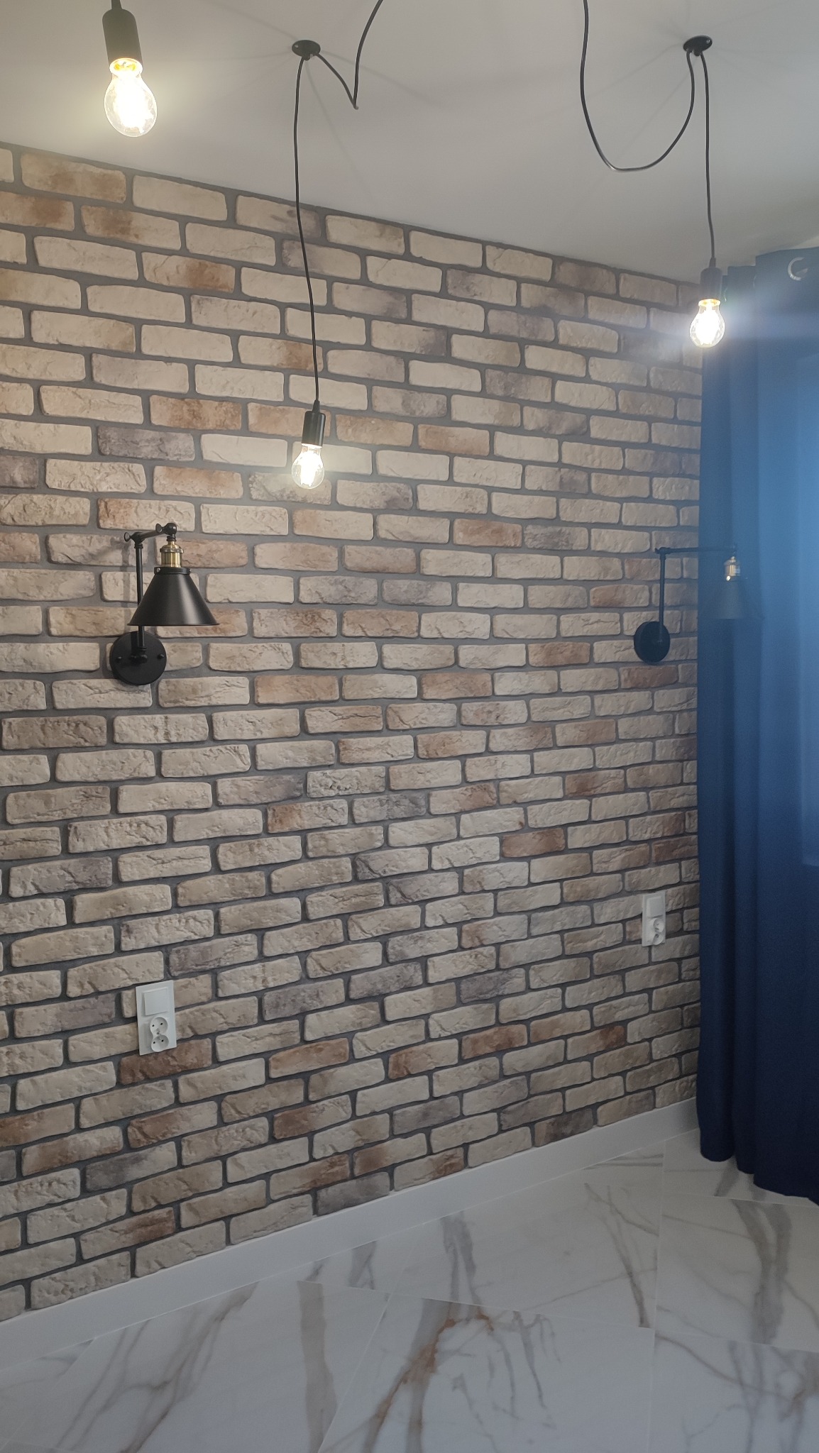 Ściana z cegły dekoracyjnej z oświetleniem: czarne kinkiety i wiszące żarówki na kablach, marmurowa podłoga, fragment zasłony.