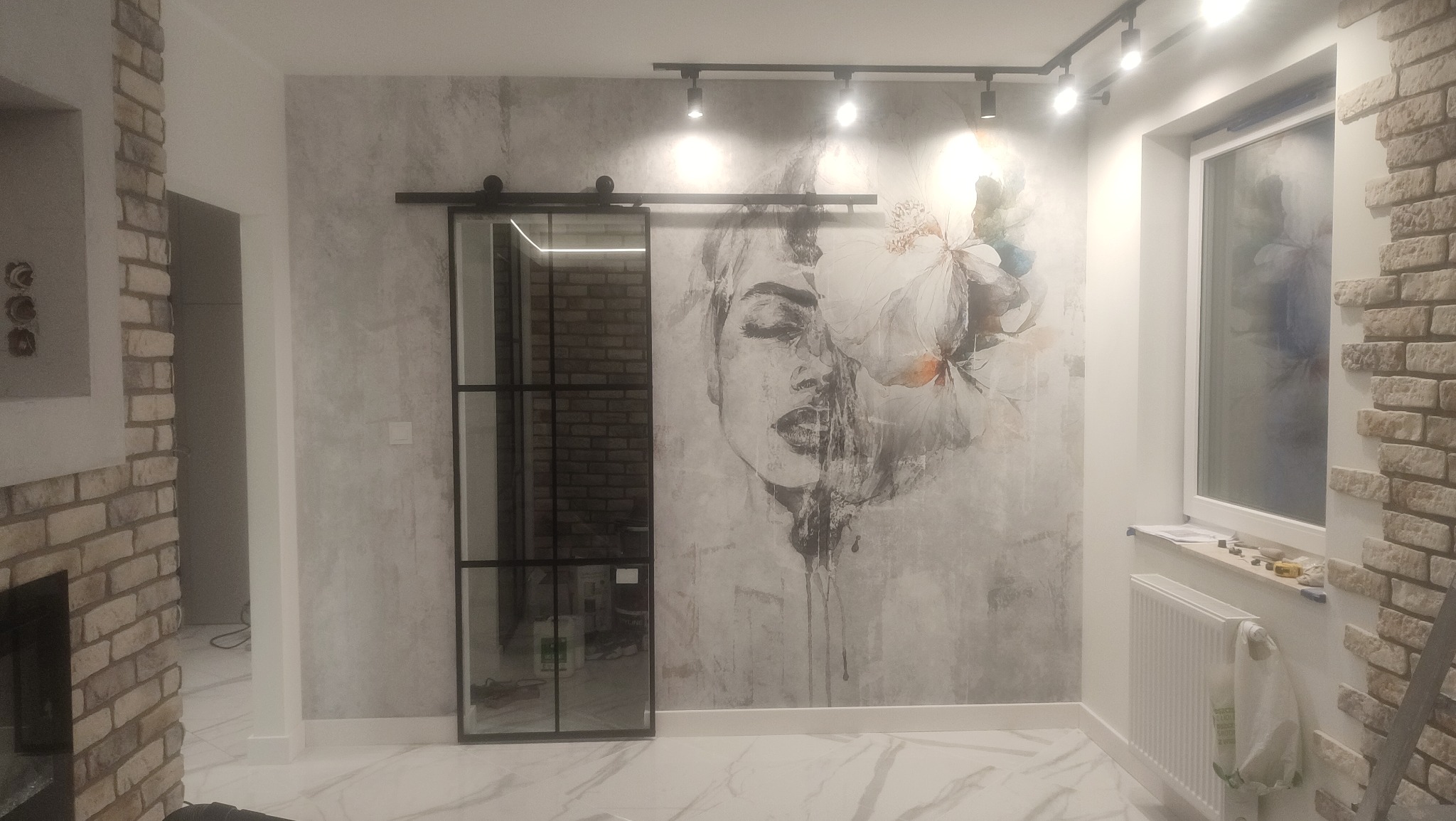 Nowoczesne wnętrze z przesuwnymi drzwiami loftowymi, marmurową podłogą, ścianą z motywem kwiatowym i ceglanym wykończeniem wnęki okiennej.