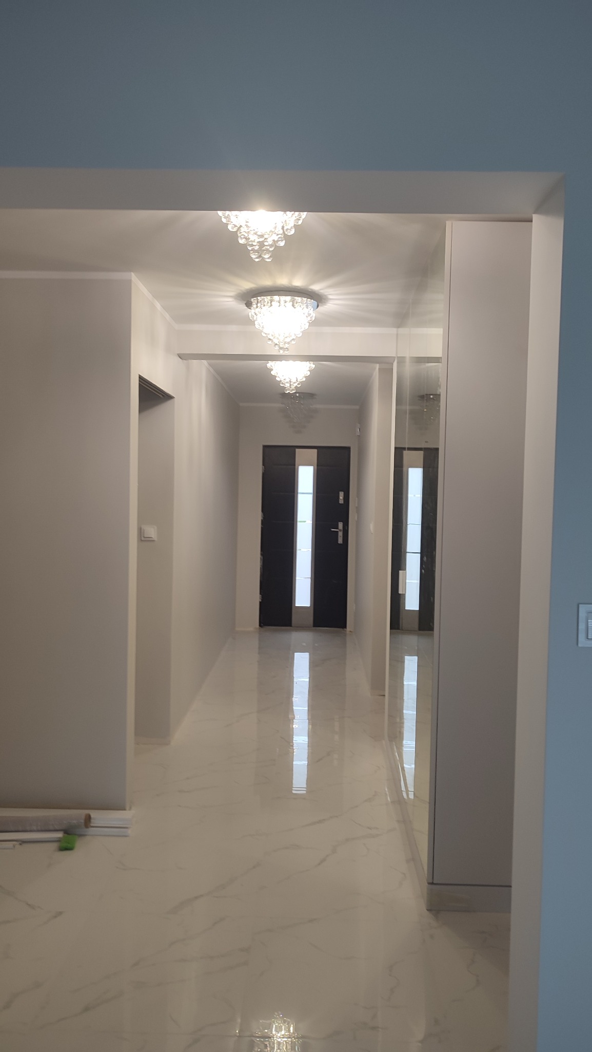 Korytarz w domu po remoncie z białymi ścianami, marmurową podłogą i trzema kryształowymi lampami na suficie, widoczna czarna futryna drzwi wejściowych.