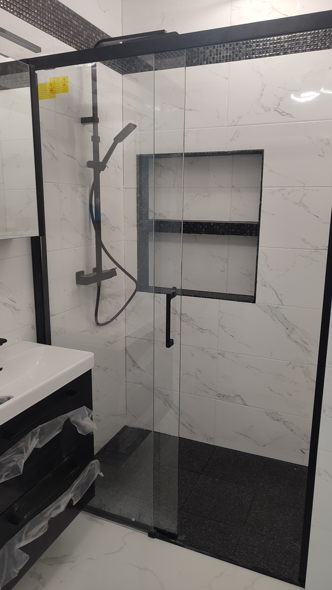 Nowoczesna kabina prysznicowa z czarnymi elementami, marmurowe płytki na ścianach, czarna podłoga w kabinie, widoczna umywalka z szafką, półka w ścianie wykończona czarną mozaiką.